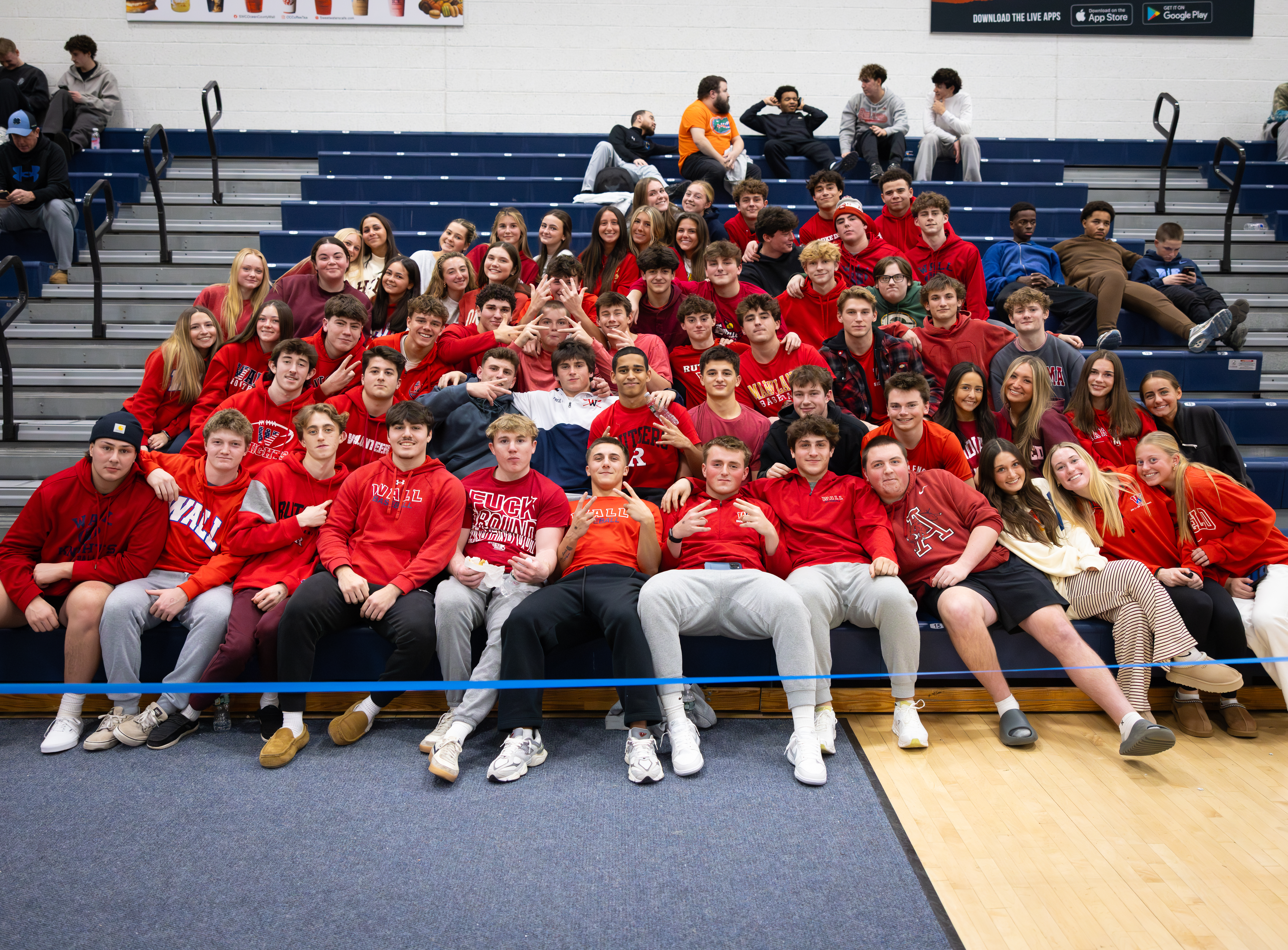 A group of young people in red shirts sit on bleachers in a gymnasium, with more spectators in the background.
