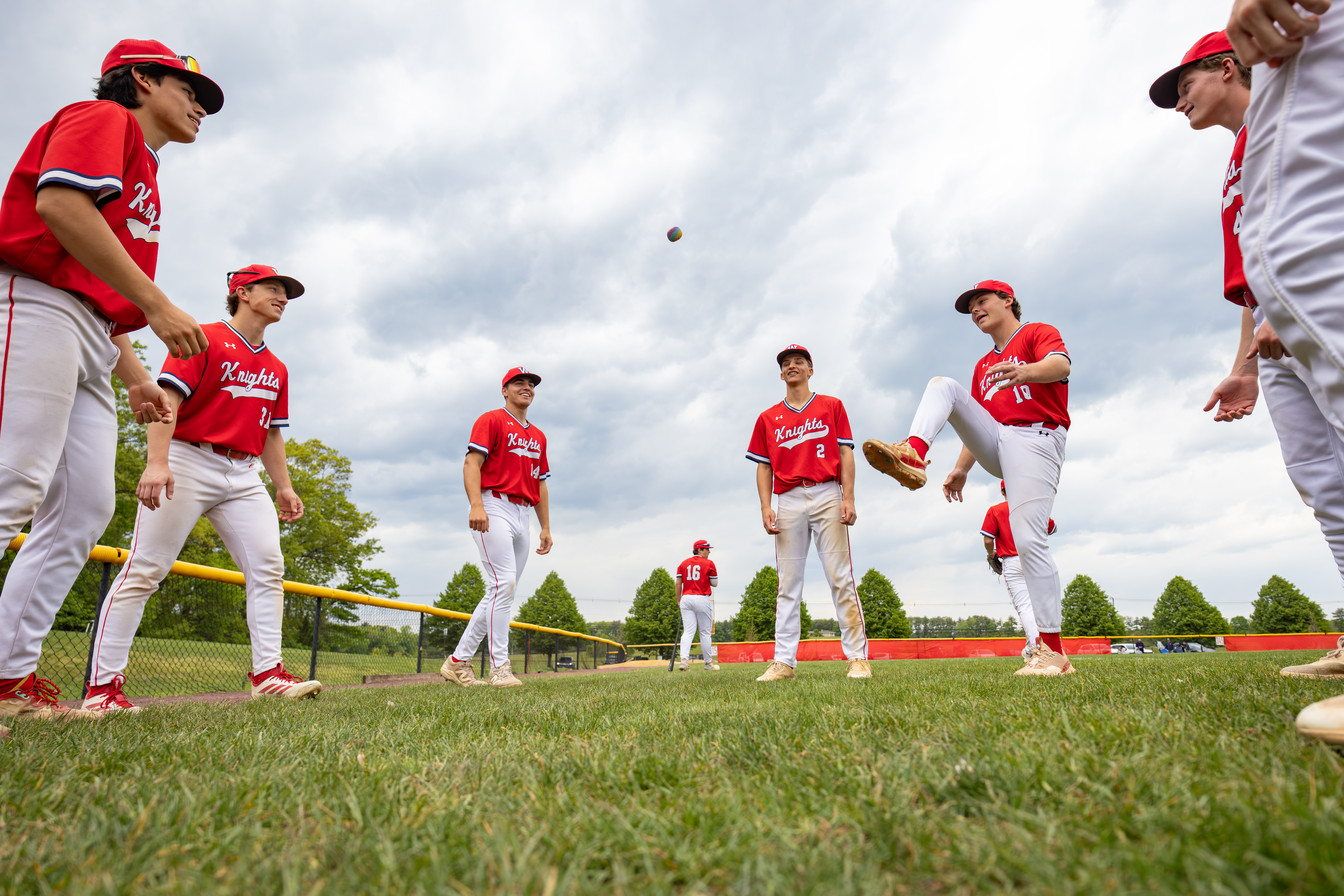 Several young baseball players in red uniforms stand on a grassy field in the foreground, while a cloudy sky and distant trees form the background.