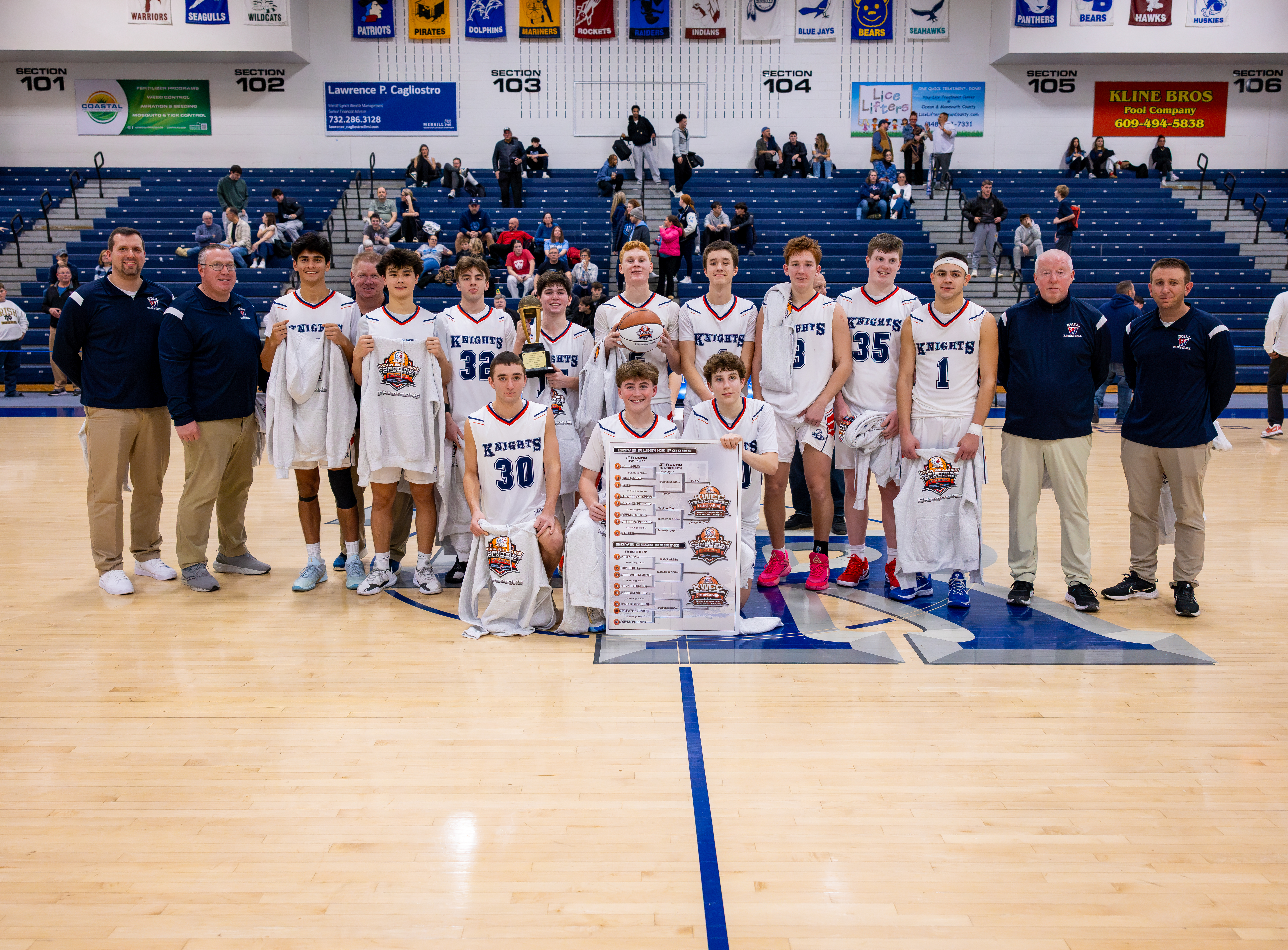 A basketball team poses for a photo on the court in front of a cheering crowd in the bleachers.