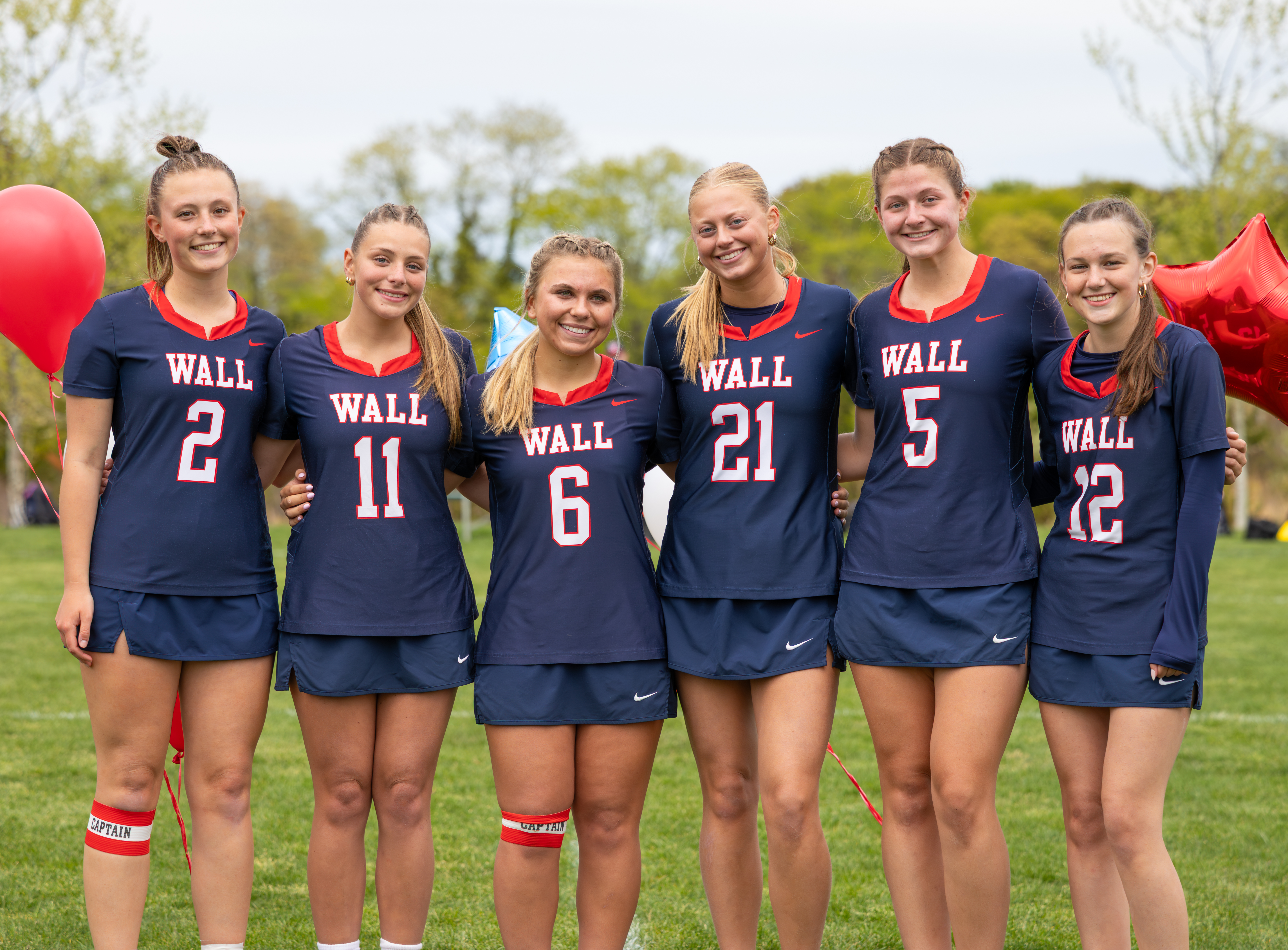 Six young women in matching navy blue lacrosse uniforms stand on a grassy field with red balloons behind them.