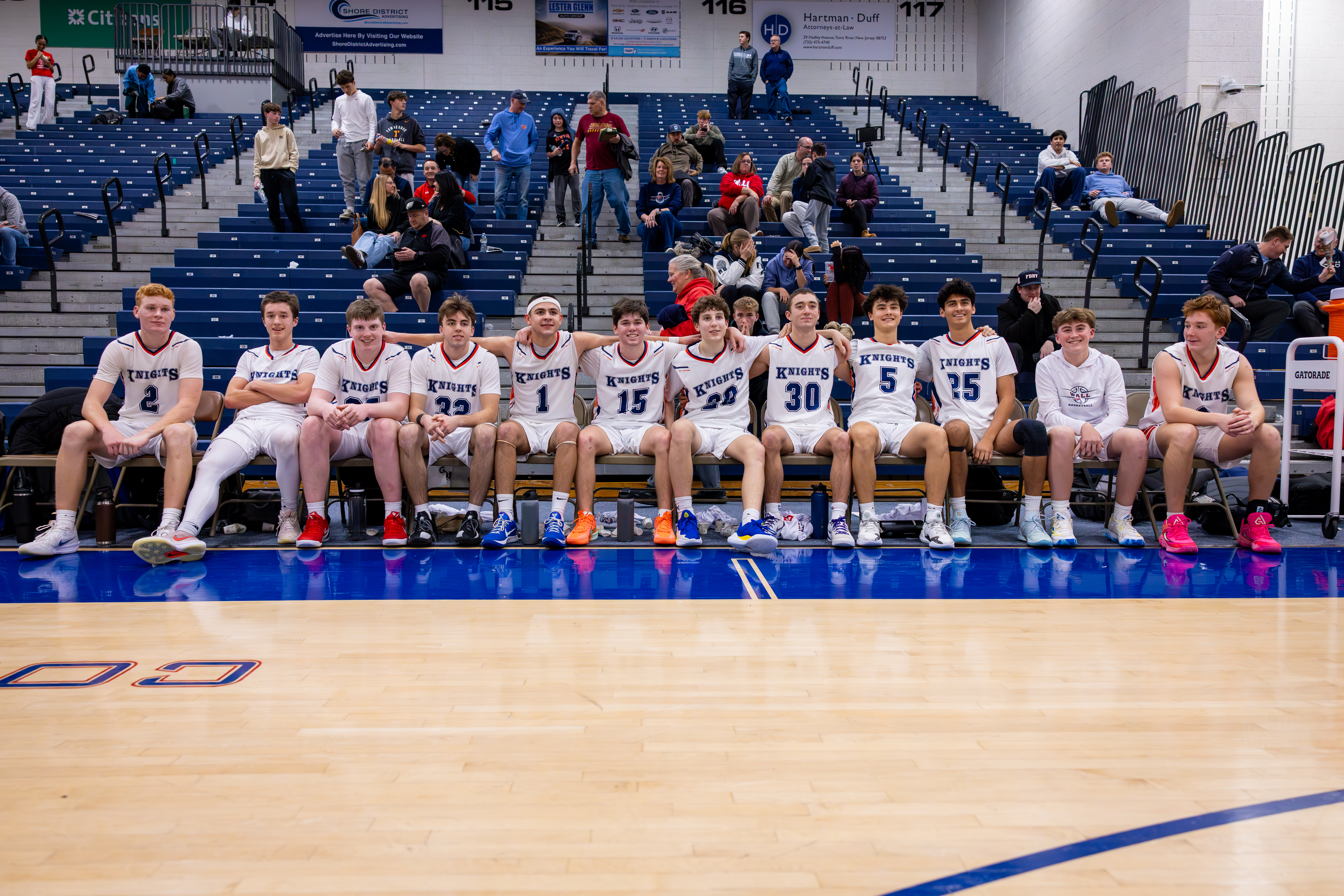 A group of young male basketball players in uniform sit on a bench in the foreground, with spectators in bleachers filling the background.