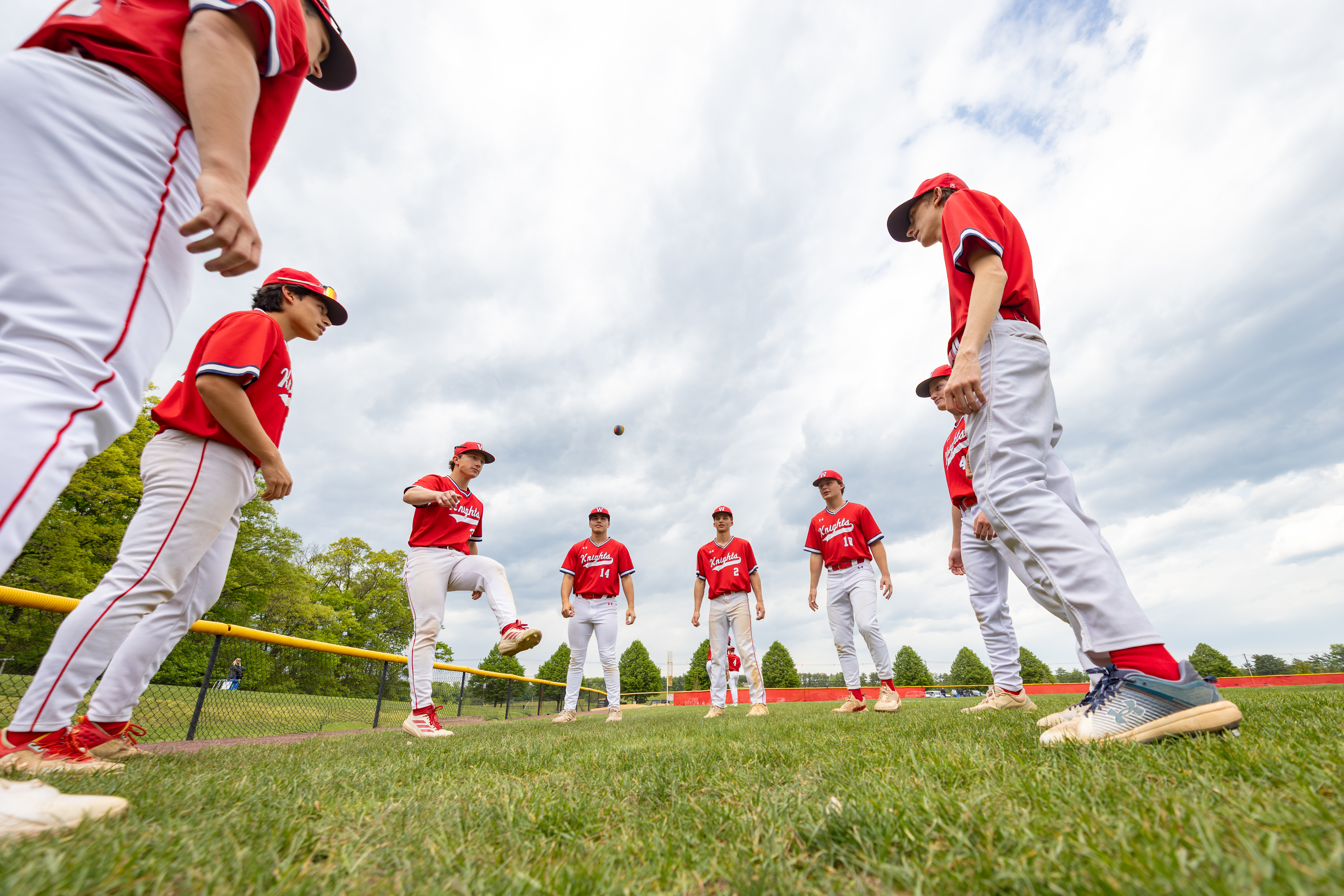 In the foreground, several baseball players in red uniforms stand on a grassy field, while in the background, a cloudy sky looms above a line of trees.