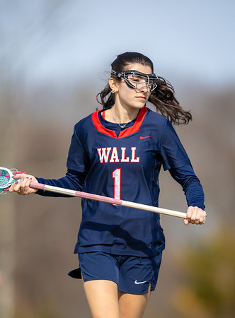 A young woman in a navy blue lacrosse uniform with "WALL 1" on the front holds her stick in the foreground, with a blurred, outdoor background.