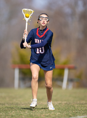 A young woman in a lacrosse uniform with a stick and ball in the foreground stands on a grassy field with blurred trees and a bench in the background.