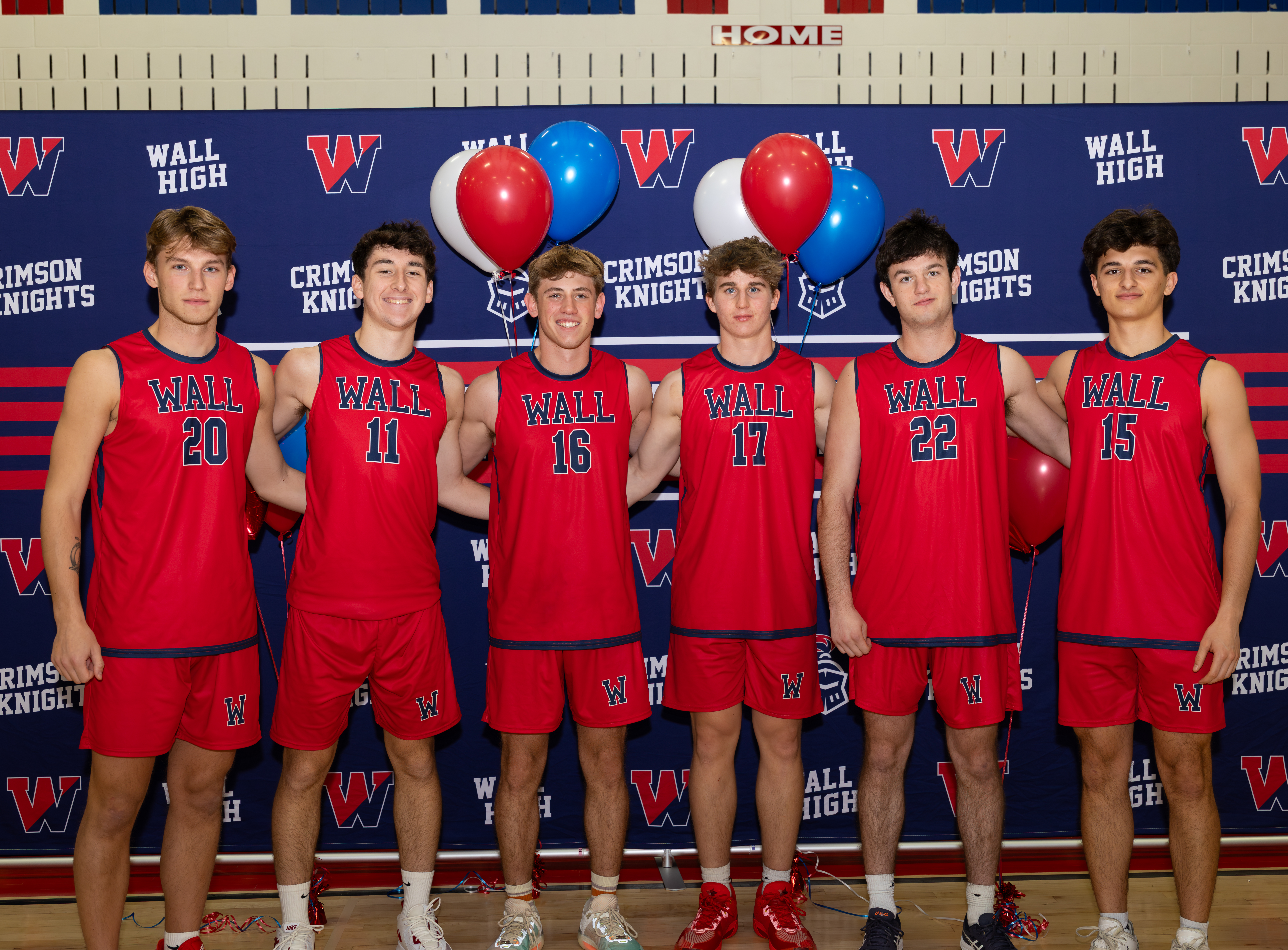 Six young men in red basketball uniforms stand in front of a blue banner with "WALL HIGH CRIMSON KNIGHTS" repeated, while balloons float above them.