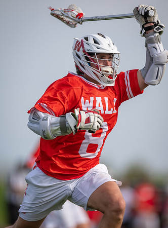 A lacrosse player in a red jersey with the number 8 on it swings his stick against a blurred background of a field and sky.