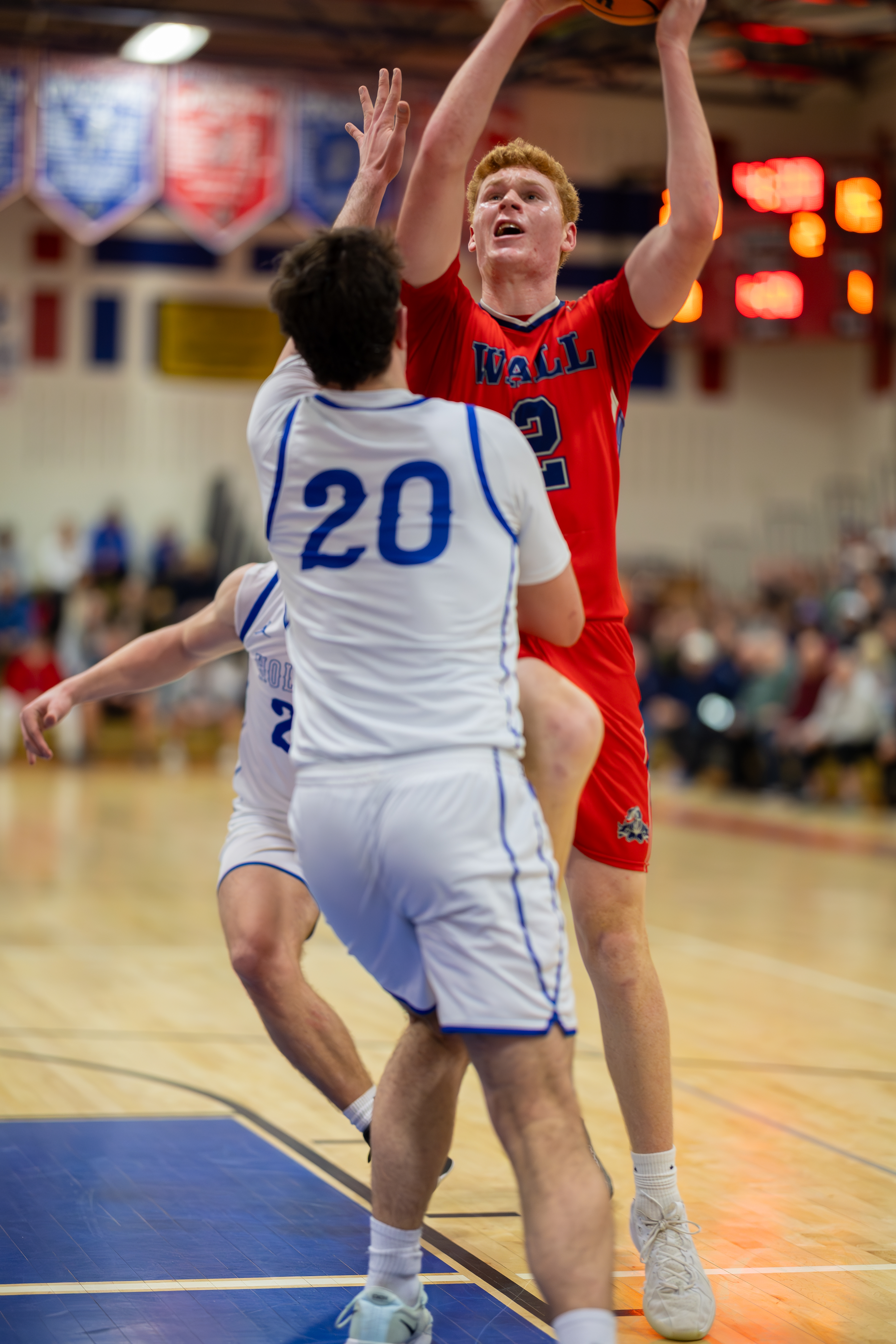 A basketball player in a red jersey with the number 2 jumps to shoot, defended by a player in a white jersey with the number 20, with a blurred crowd and scoreboard in the background.