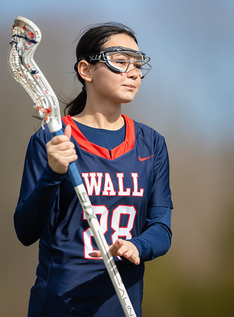 A young girl in a navy blue lacrosse jersey with "WALL 28" on it holds a lacrosse stick in the foreground, with a blurred background of trees and sky.