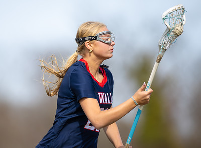 A blonde female lacrosse player in a navy blue jersey with the number 22 on it, wearing goggles and holding her stick, is captured in profile against a blurred background of trees and sky.