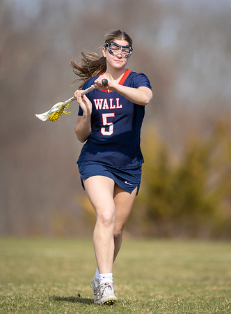 A young woman in a navy blue lacrosse uniform with the number 5 on it runs across a grassy field, holding her stick, with a blurred background of trees.