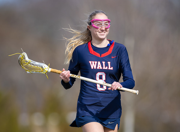 A smiling young woman in a lacrosse uniform holds her stick in the foreground, with a blurred background of trees and sky.