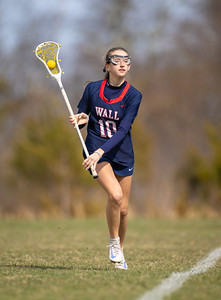 A young woman in a lacrosse uniform runs on a grassy field with a stick and ball, while trees blur in the background.