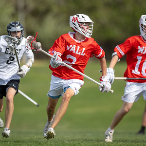 Three young men in lacrosse gear run across a grassy field, with one in the foreground wearing a red jersey and white helmet.