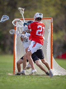 Two lacrosse players in red and white uniforms are in the foreground, battling for the ball in front of a net, with a blurred green field and trees in the background.