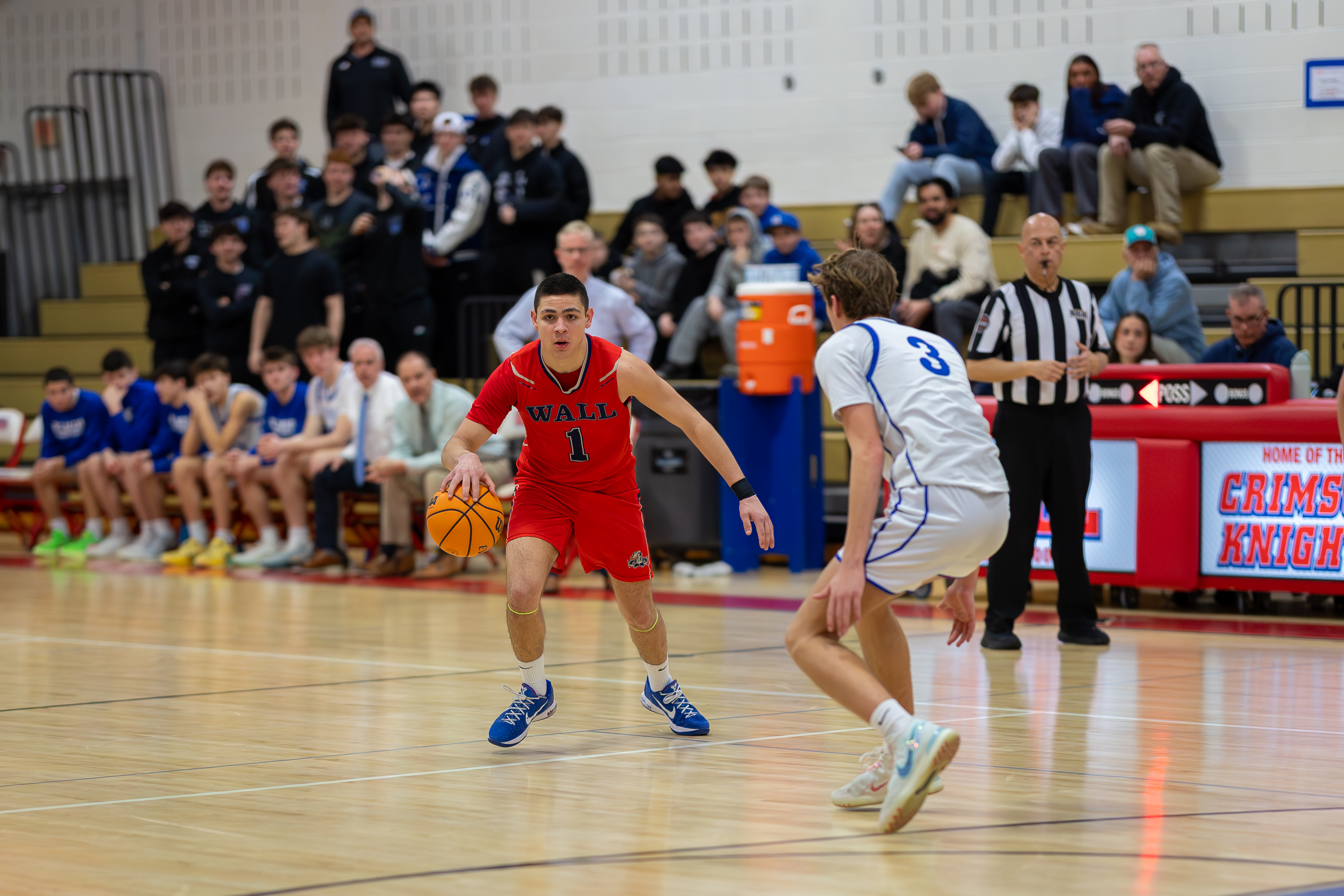 A basketball player in a red jersey dribbles the ball on a court while spectators watch from bleachers in the background.