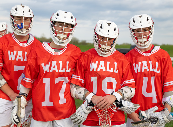 Four young men in orange lacrosse jerseys and white helmets stand in the foreground, while a blurred green field and sky form the background.
