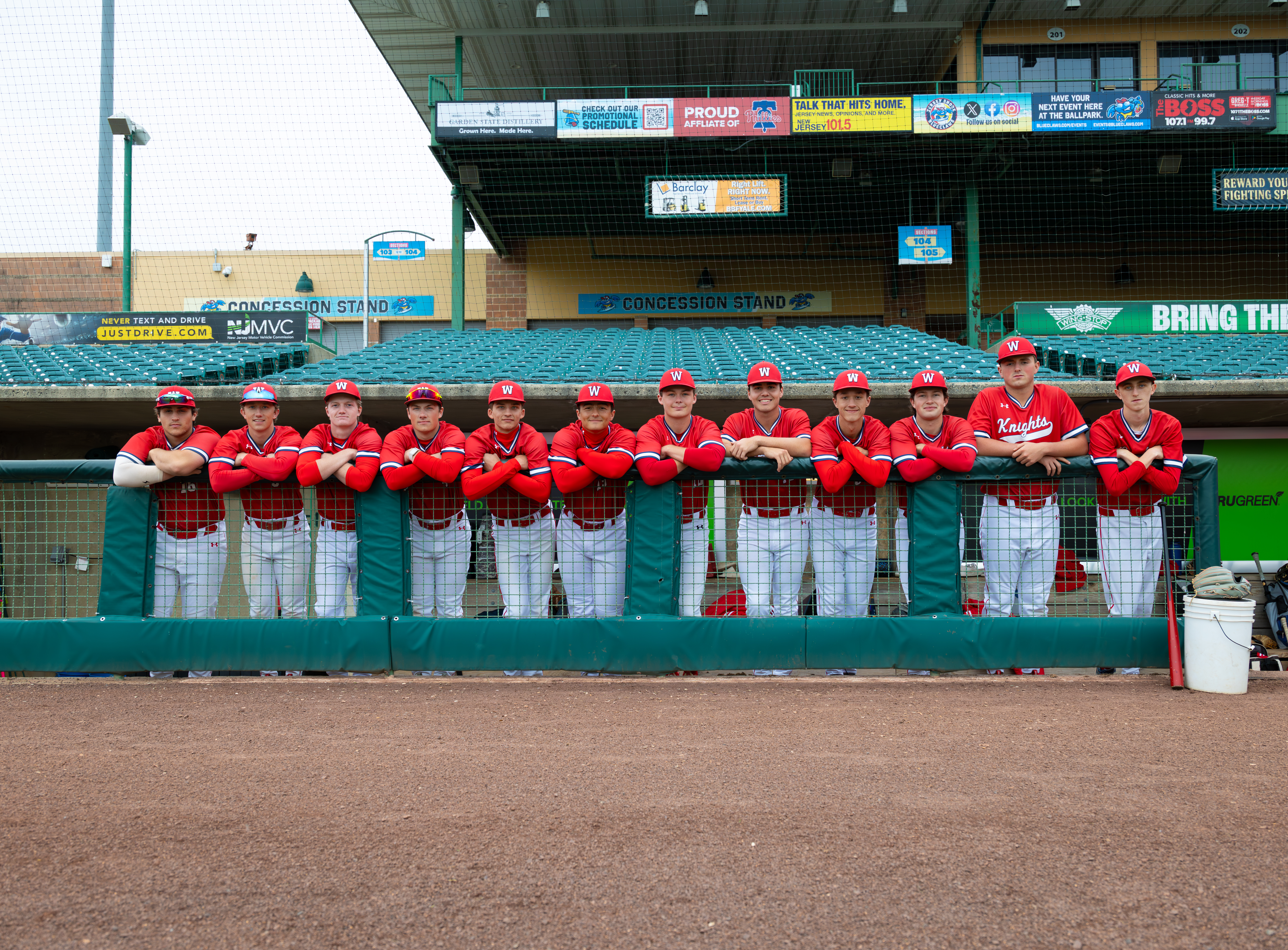 A baseball team of twelve boys in red and white uniforms stand behind a green dugout railing in front of a stadium with concession stands and advertising in the background.