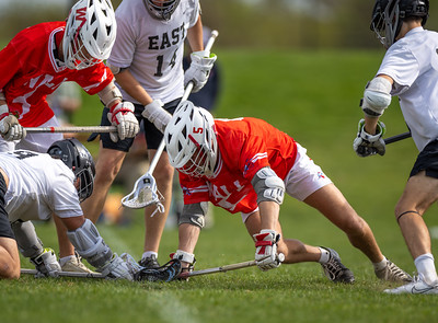 Lacrosse players in red and white uniforms scramble for the ball on a grassy field with a blurred background of trees and sky.