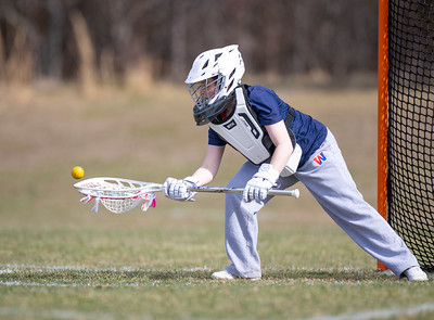 A lacrosse goalie in the foreground lunges to catch a ball, with a net and blurred trees in the background.