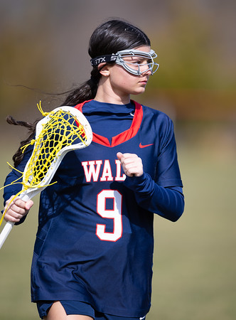 A young woman in a navy blue lacrosse uniform with the number 9 on it runs on a grassy field, holding her stick, with a blurred background of trees and sky.