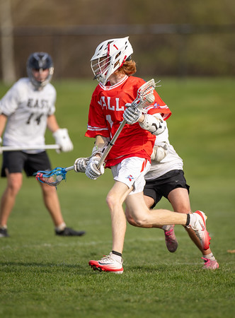 A young lacrosse player in a red jersey runs with the ball on a grassy field, while another player in a white jersey is visible in the blurred background.