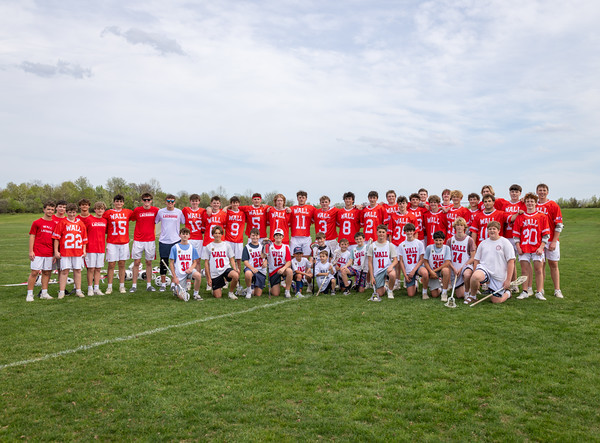 A large group of young lacrosse players in red jerseys pose on a grassy field with a cloudy sky in the background.