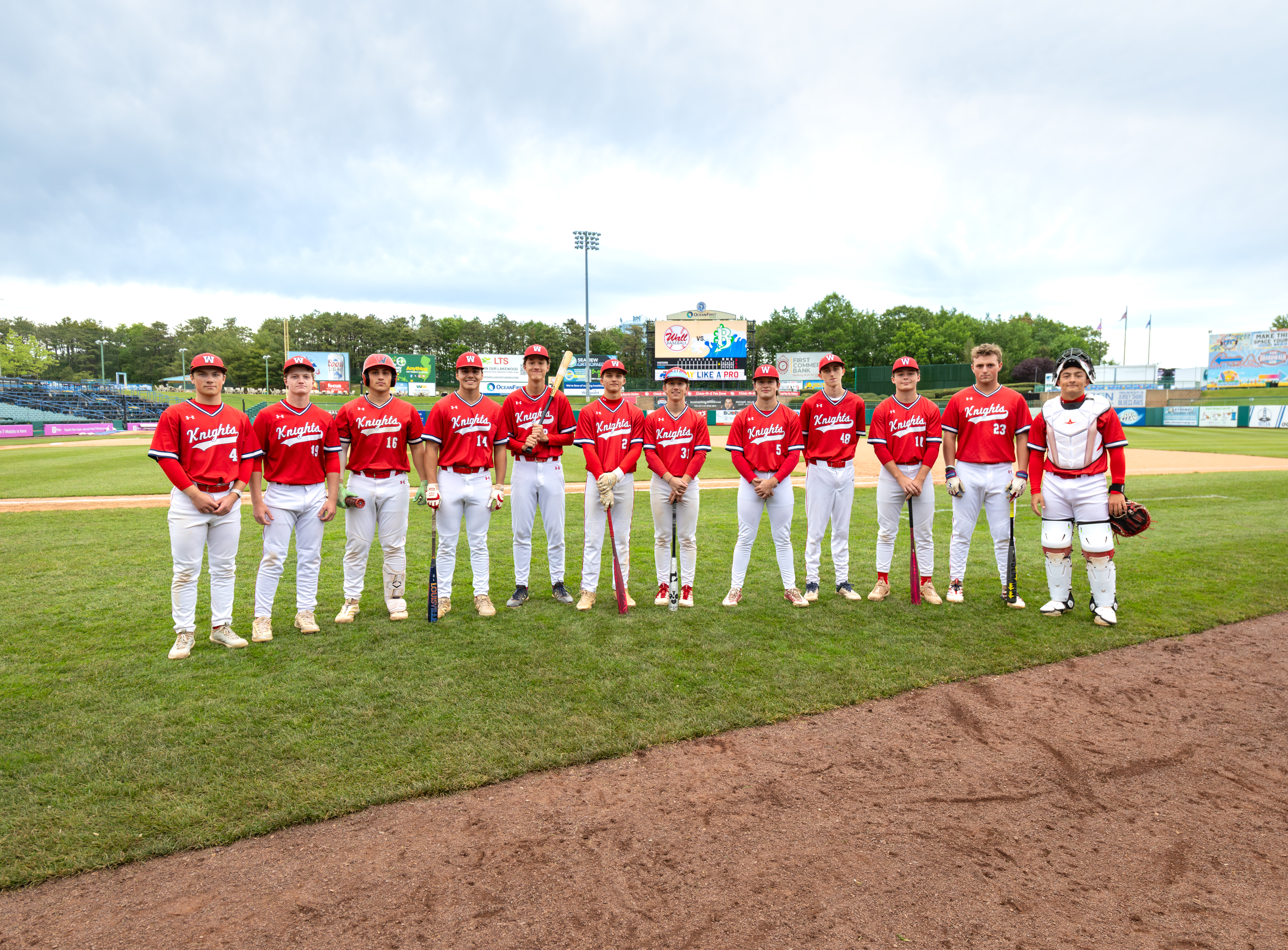 A baseball team in red uniforms stands on a grassy field in front of a stadium under a cloudy sky.