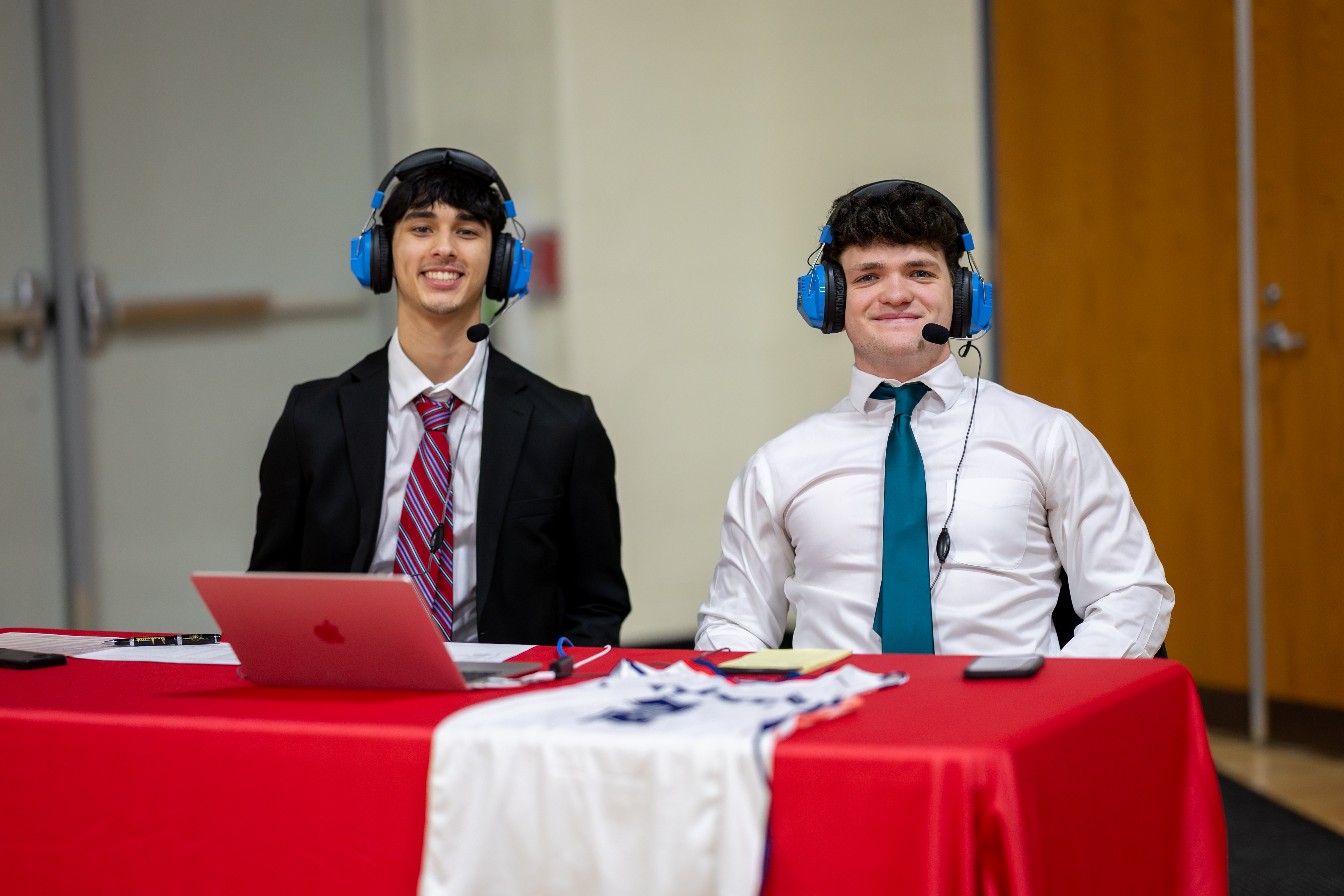 Two young men in suits and headsets smile from behind a red-covered table with a laptop and a jersey, in front of a wooden gymnasium wall.
