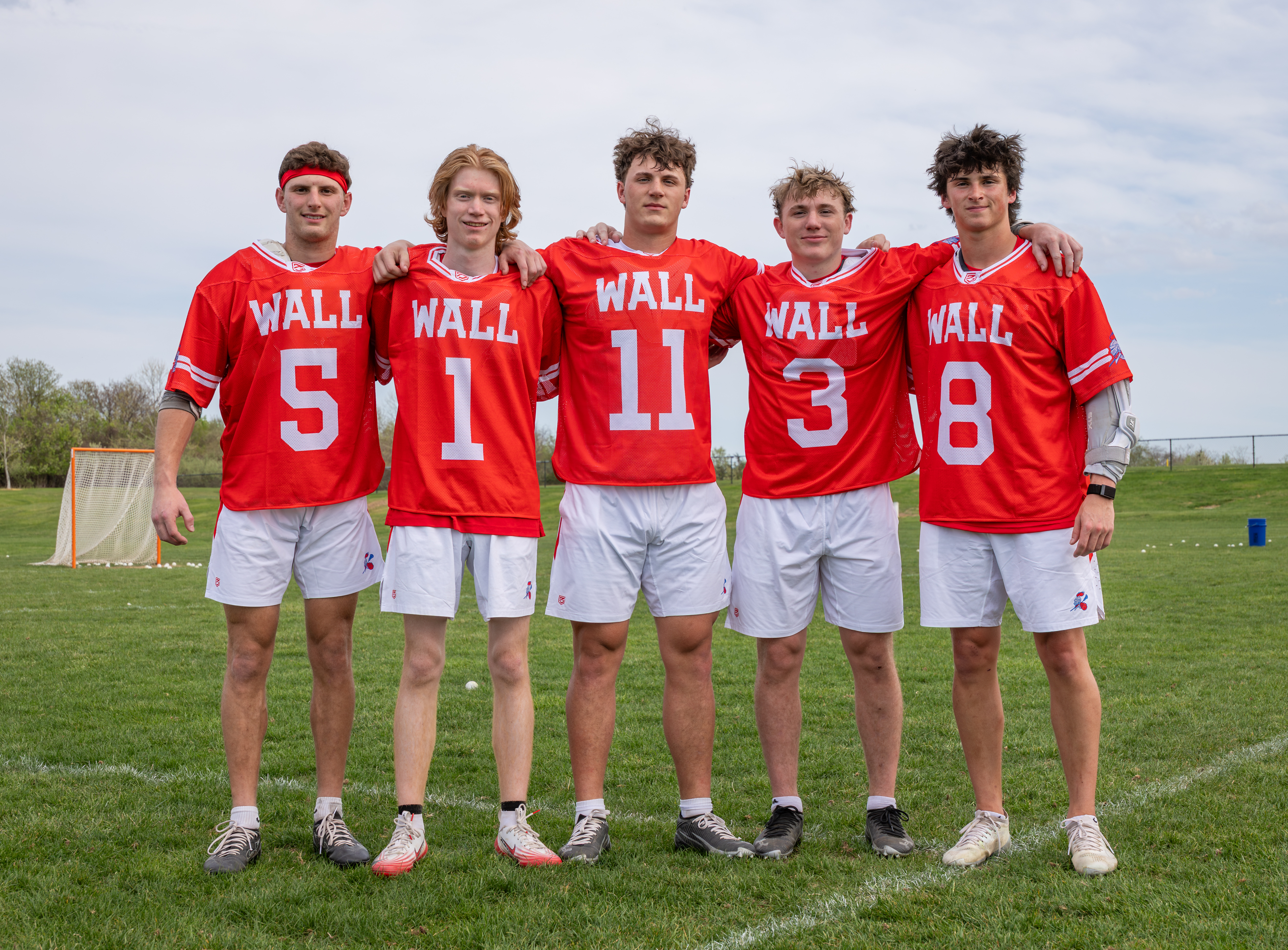 Five young men in red "WALL" jerseys and white shorts stand on a grassy field with a lacrosse goal in the background.