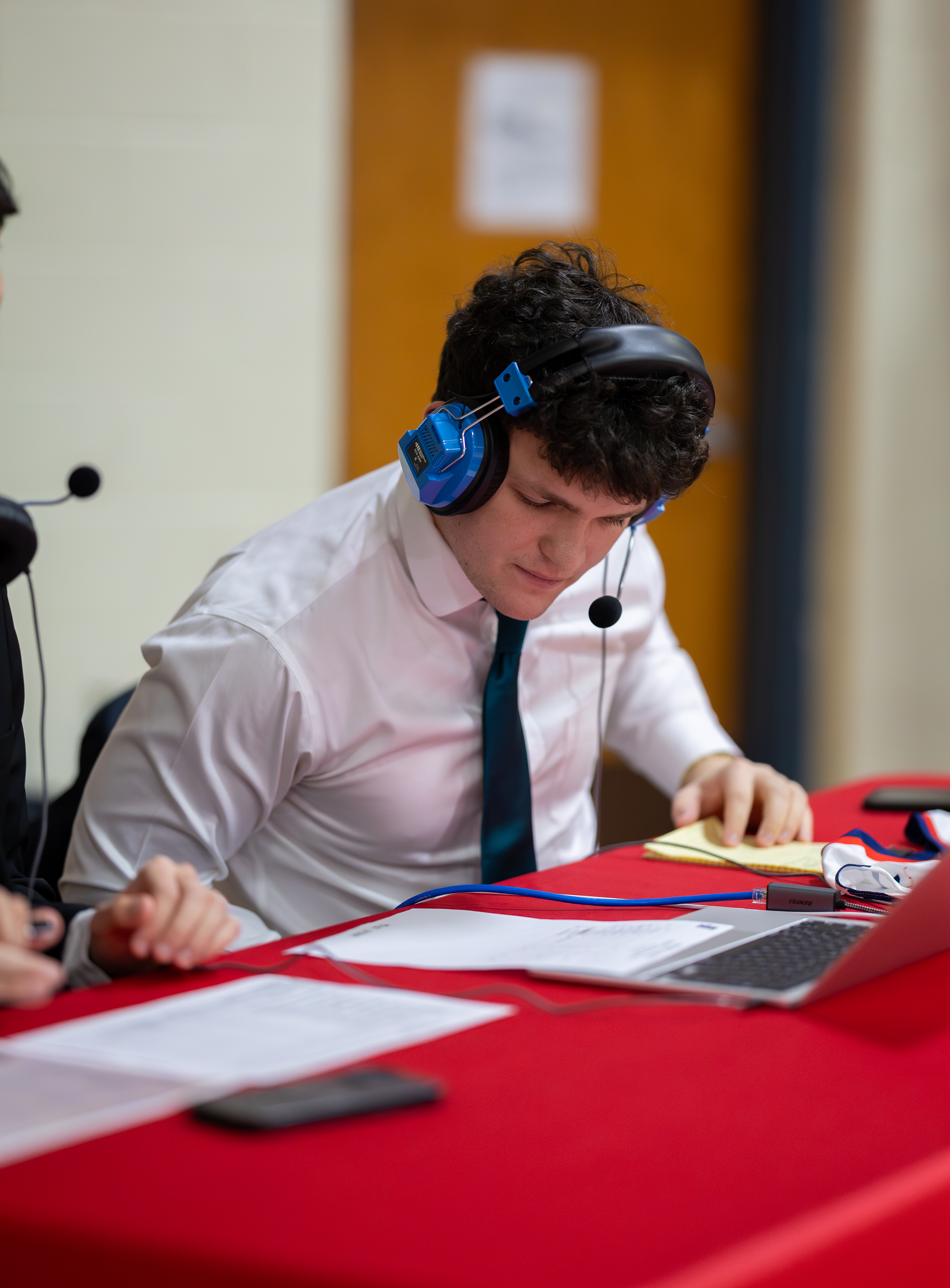 A young man in a white shirt and tie, wearing blue headphones, looks down at papers and a laptop on a red table, with a blurred background of a room.