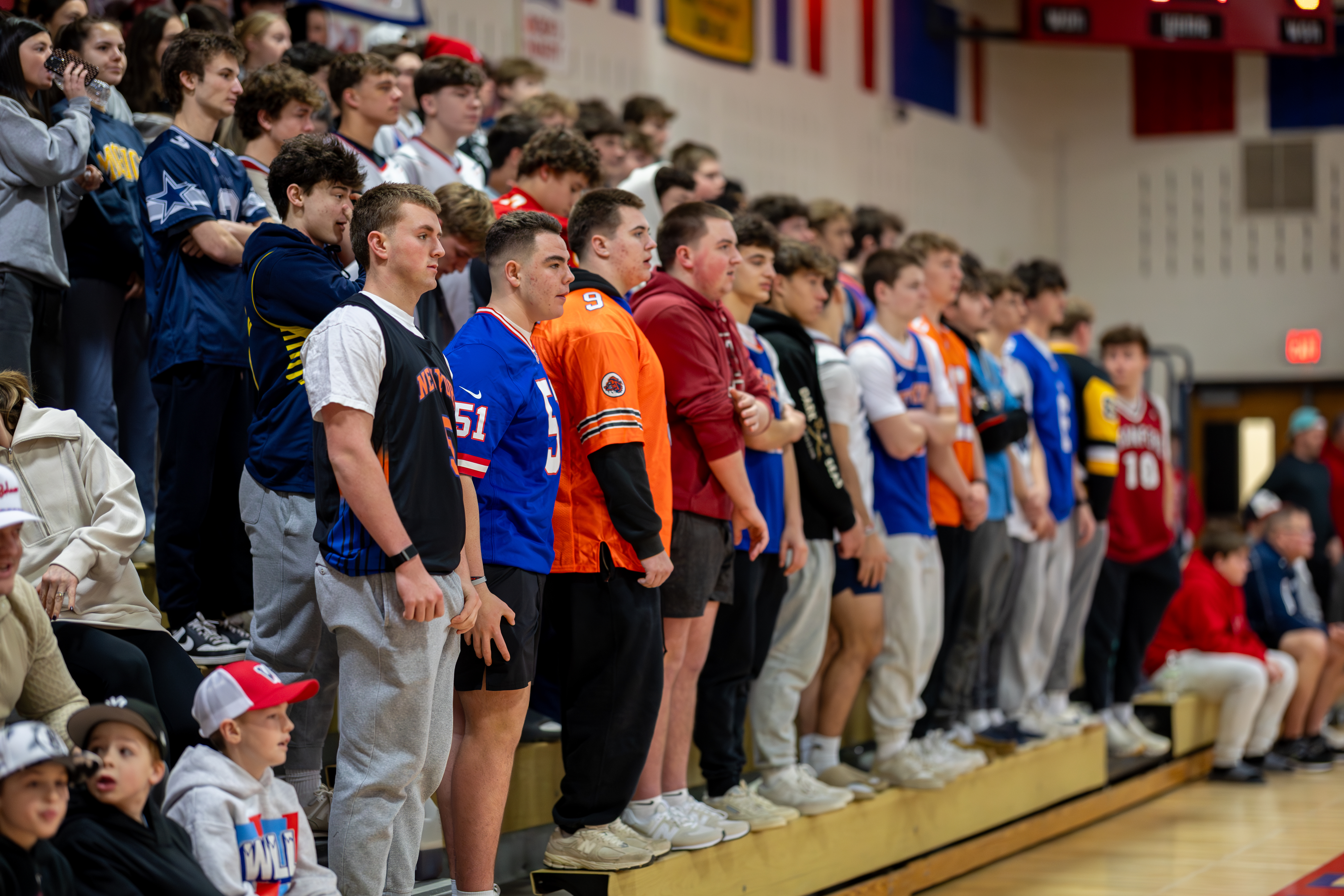 A crowd of spectators, many wearing jerseys, stands and sits in bleachers in a gymnasium with flags hanging in the background.