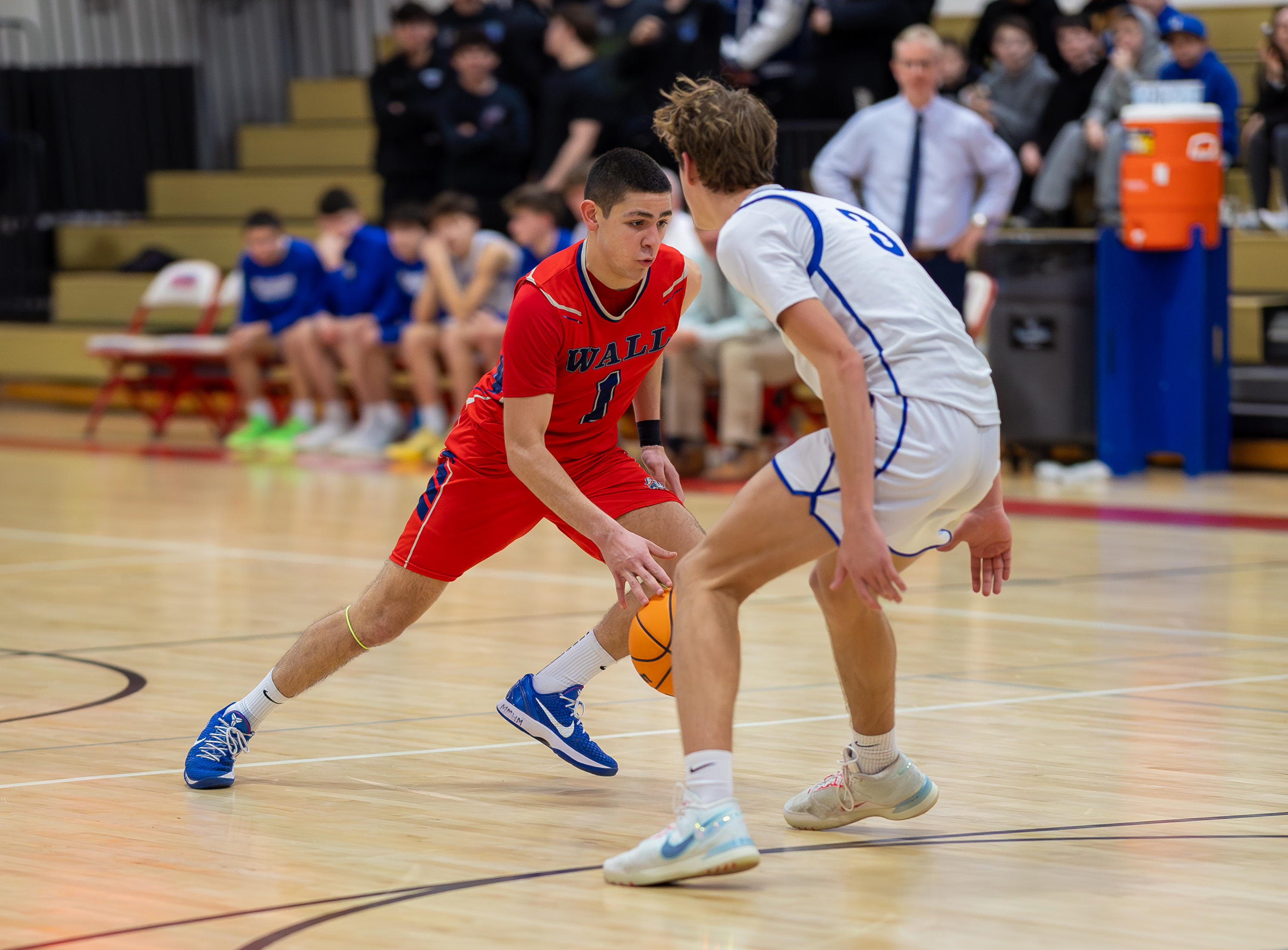 Two young basketball players in red and white uniforms dribble and defend on a wooden court, with spectators in bleachers visible in the blurred background.