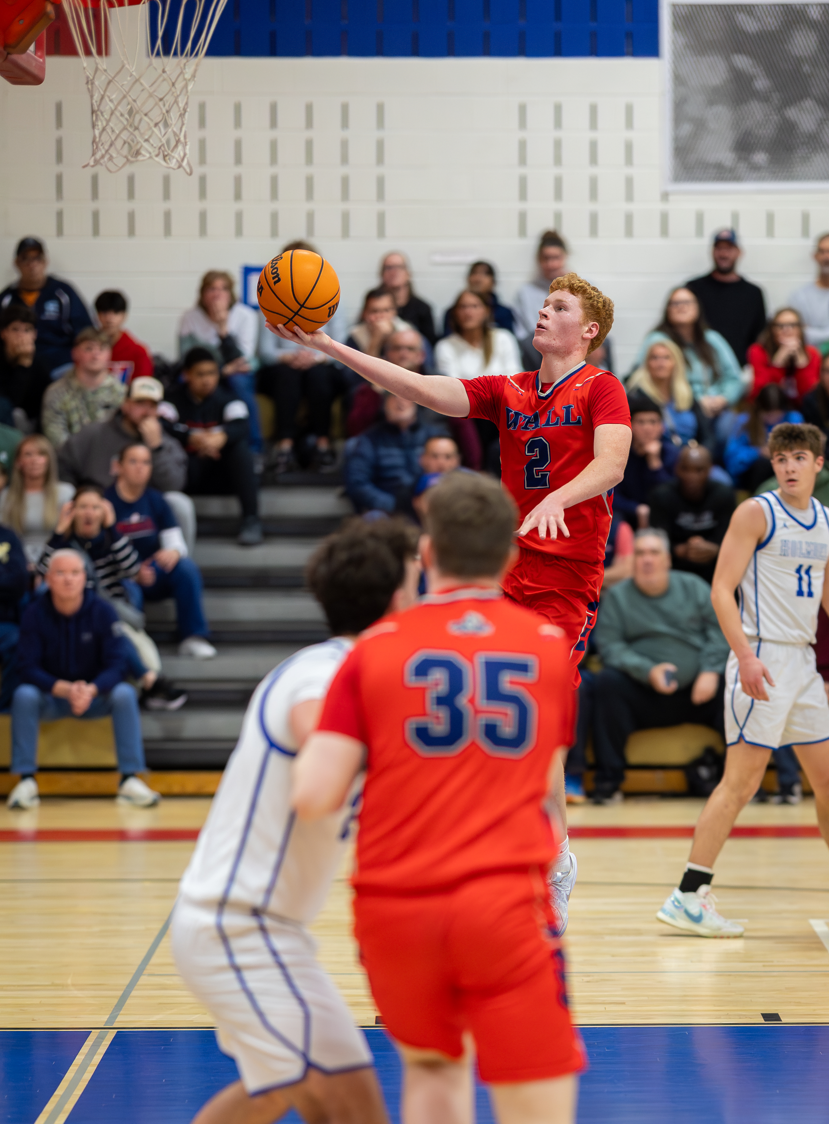 A basketball player in a red jersey with the number 2 on it shoots the ball in the foreground, while spectators fill the bleachers in the background.