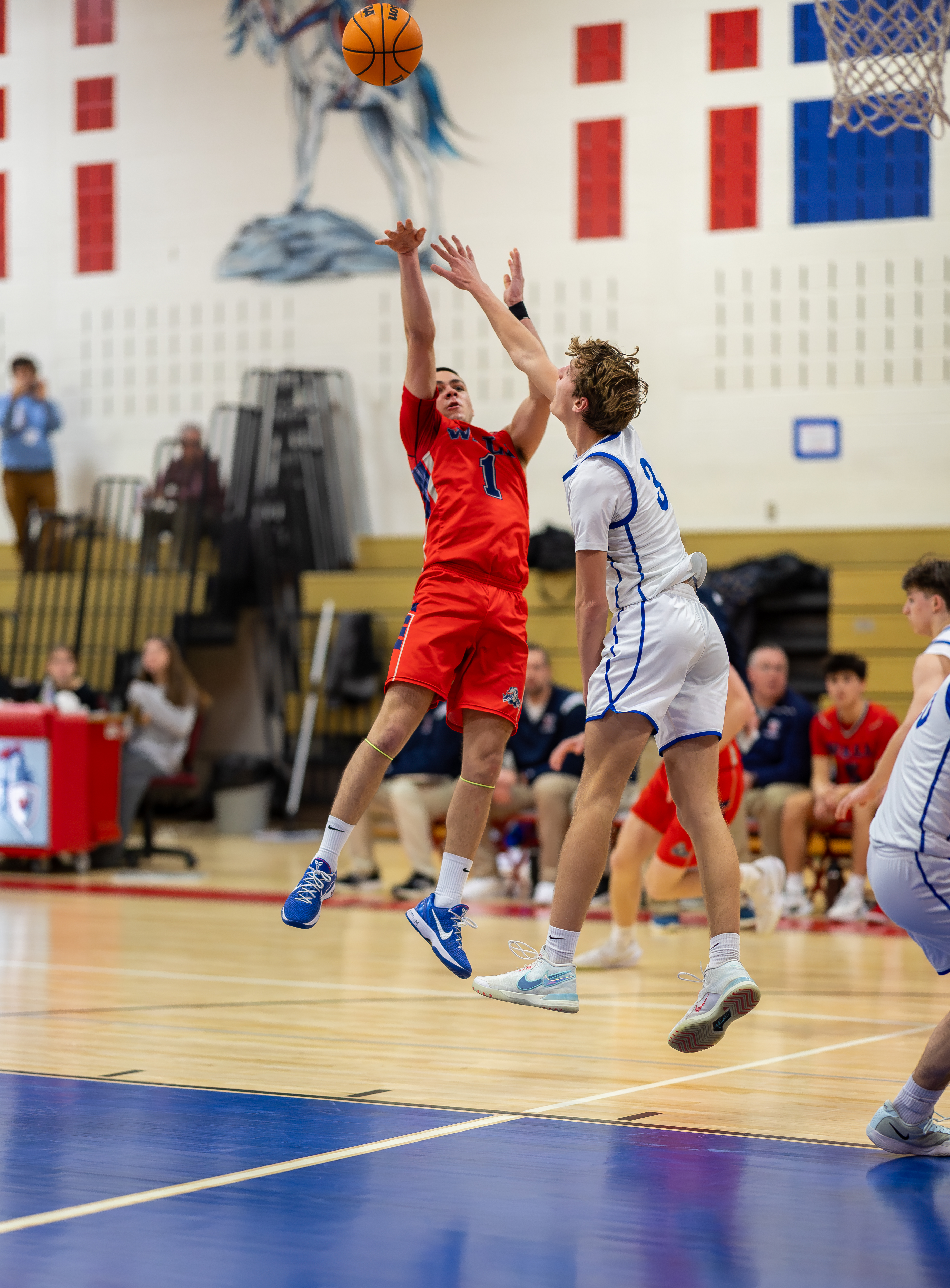 A basketball player in a red uniform shoots a ball while defended by a player in a white uniform, with spectators and a mural of a horse visible in the background.