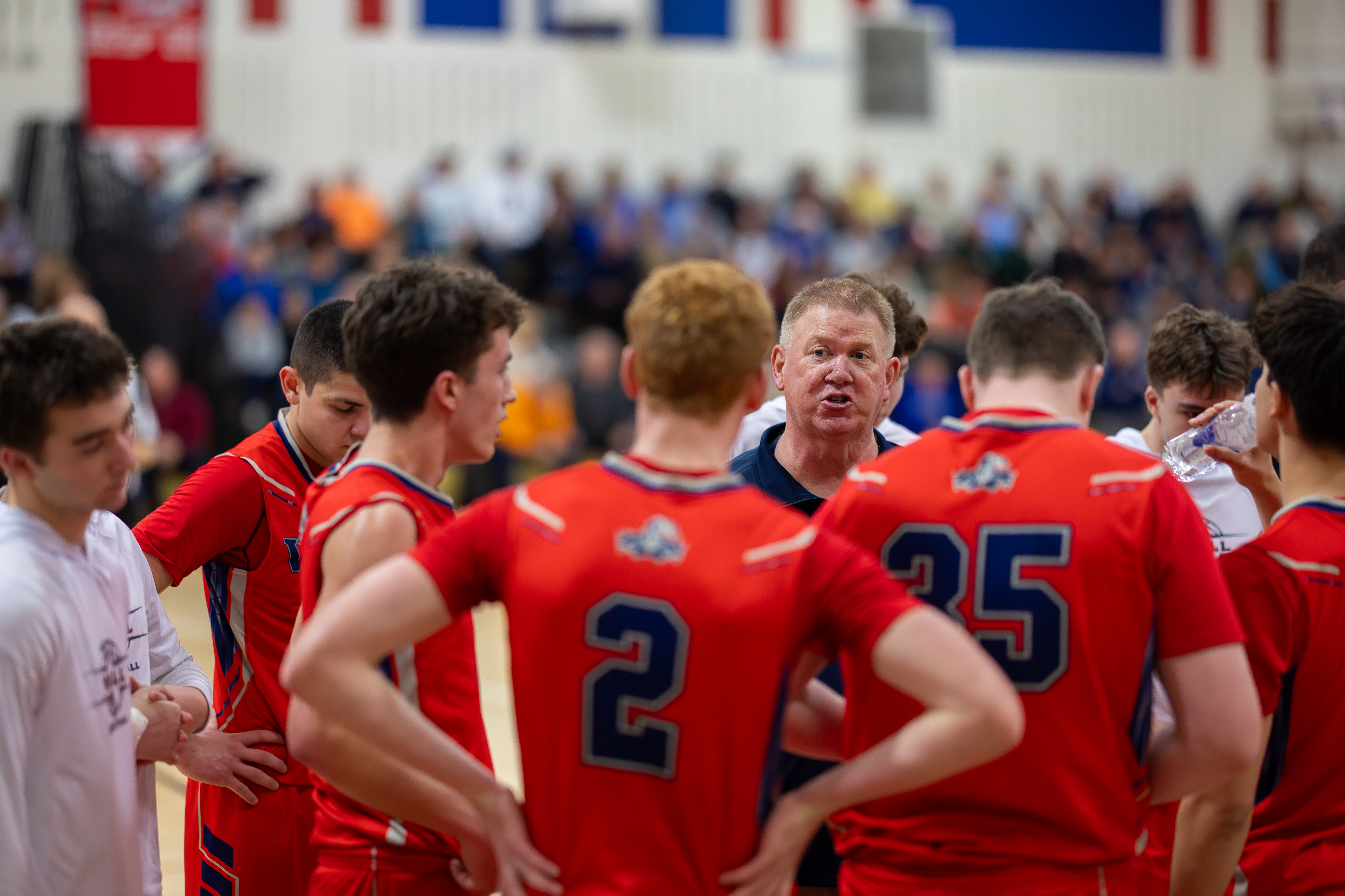 A basketball coach in the foreground addresses his team in red jerseys, while a blurred crowd watches from the background.