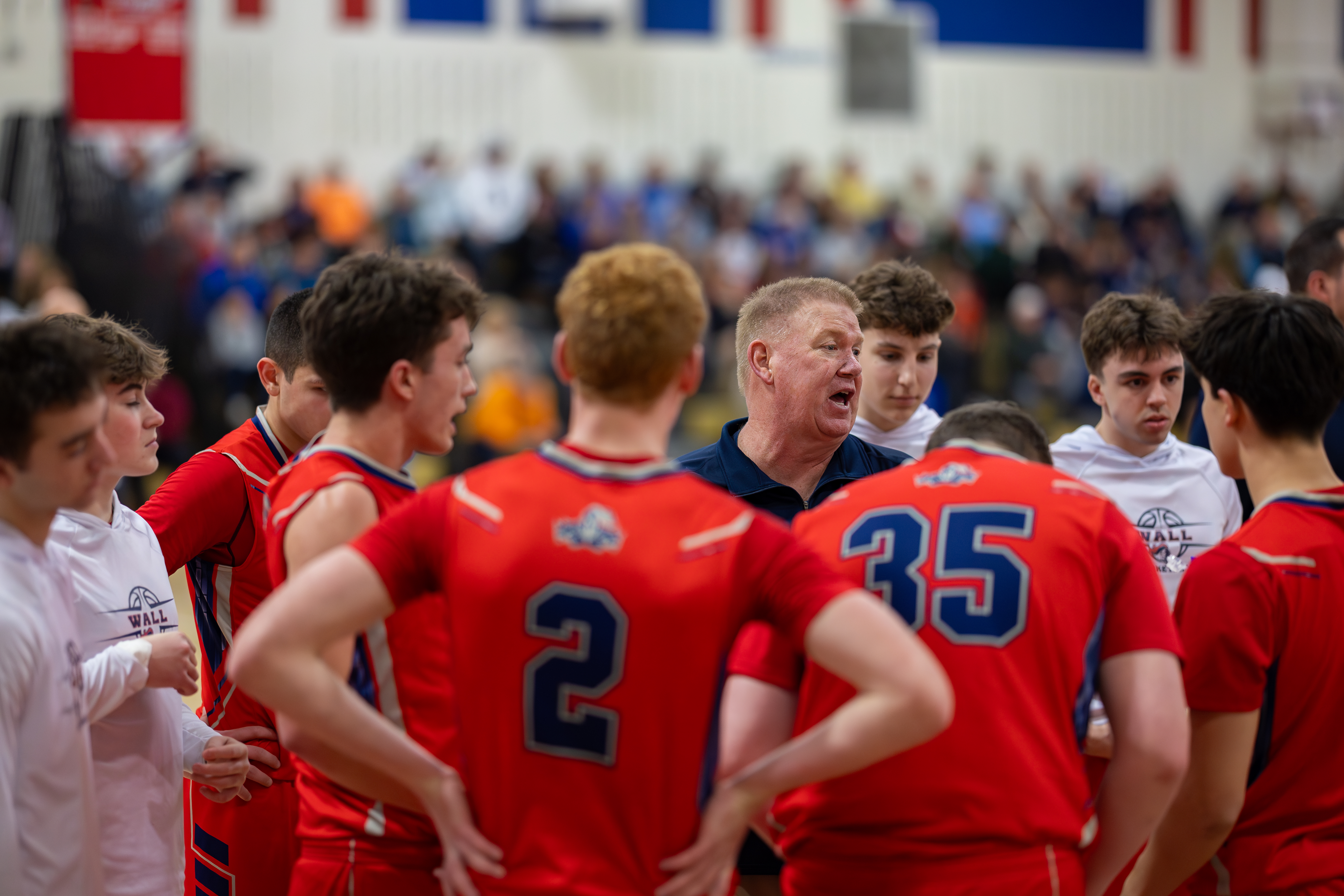 A basketball coach in a blue shirt gestures to a huddle of players in red jerseys, with a blurred crowd watching in the background.