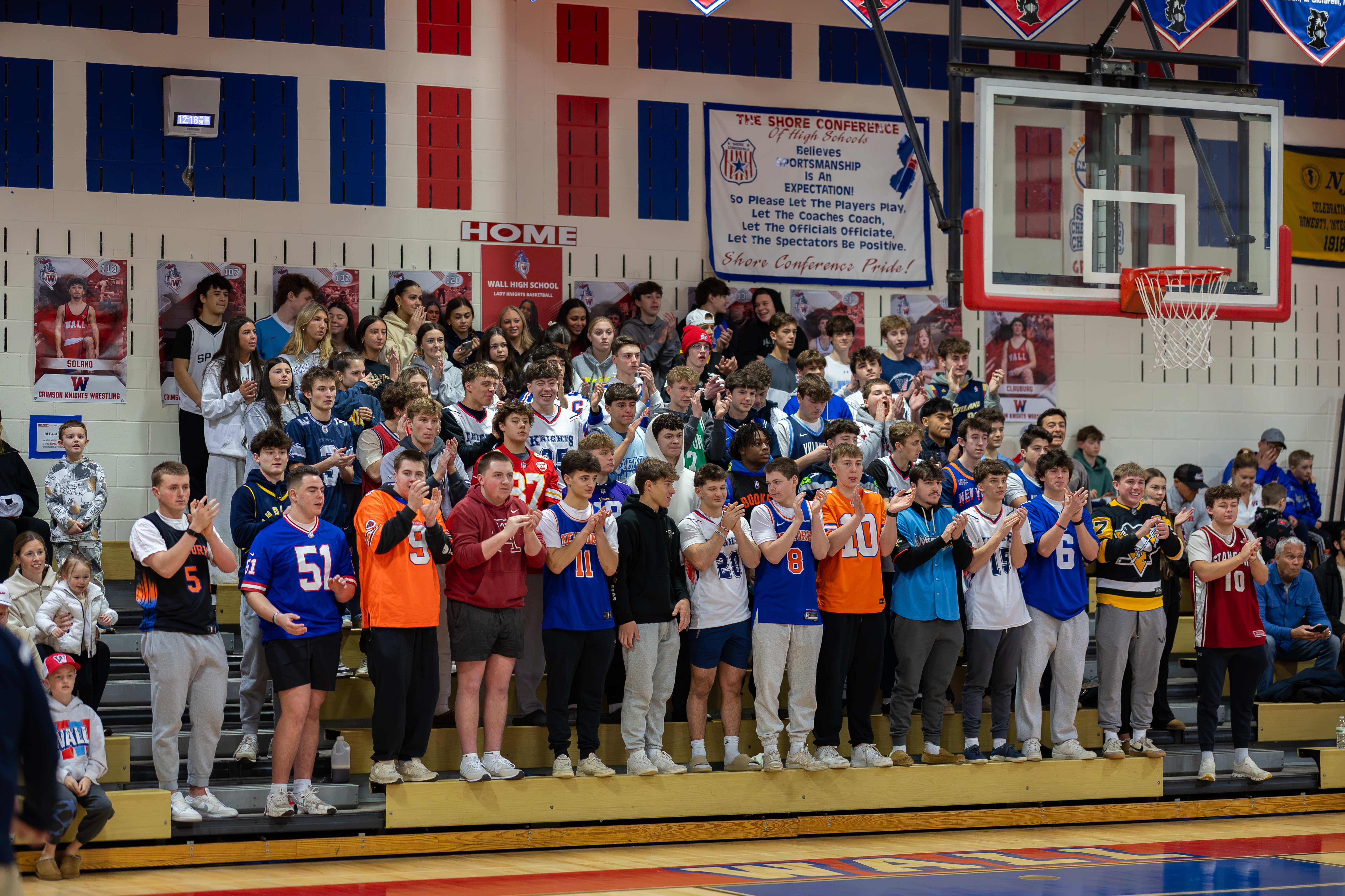 A crowd of students in athletic jerseys cheers from bleachers in a gymnasium, with a basketball hoop visible in the background.