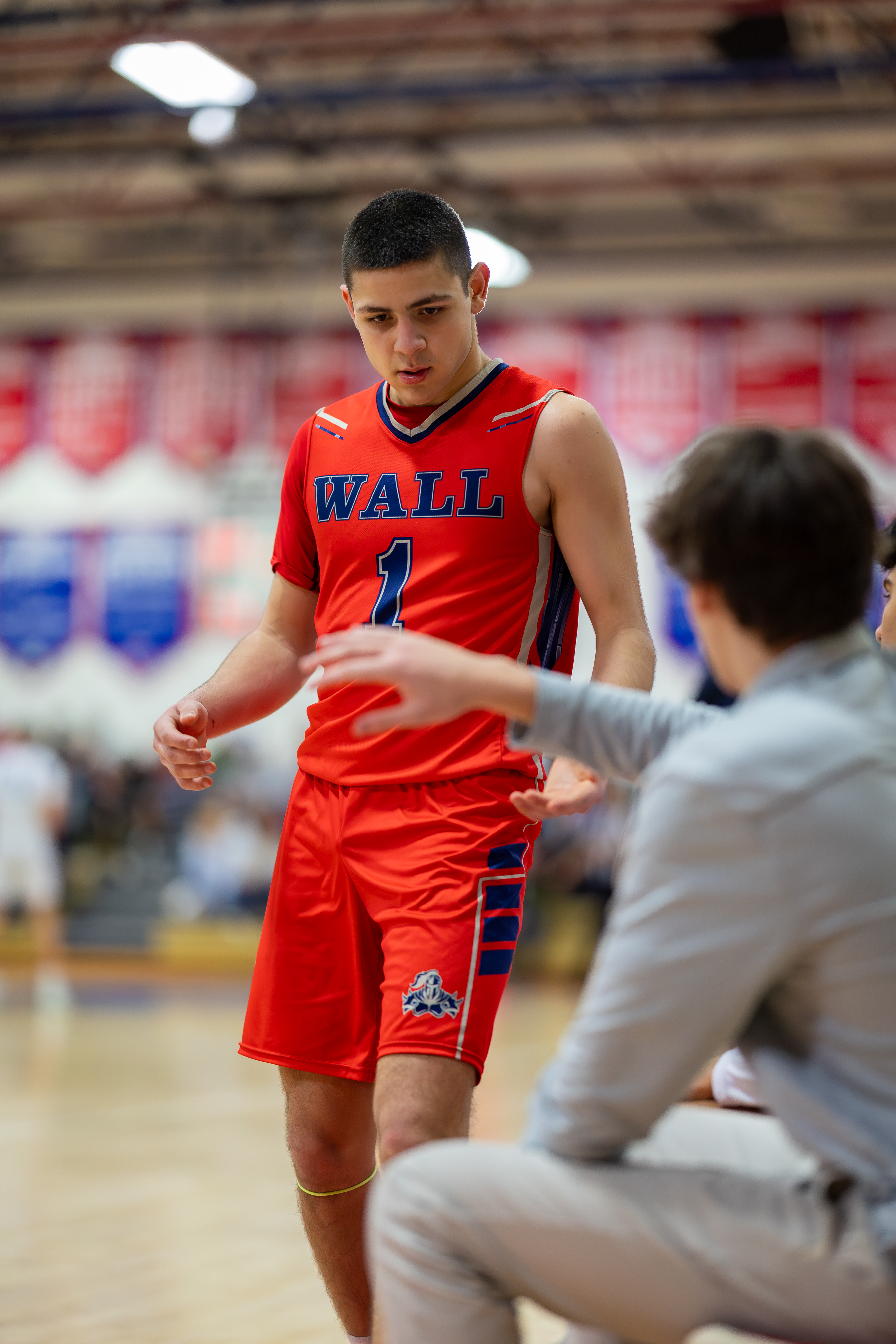 A young basketball player in a red jersey with the number 1 stands on a court, while another player in a gray shirt is in the foreground, with blurred spectators and banners in the background.