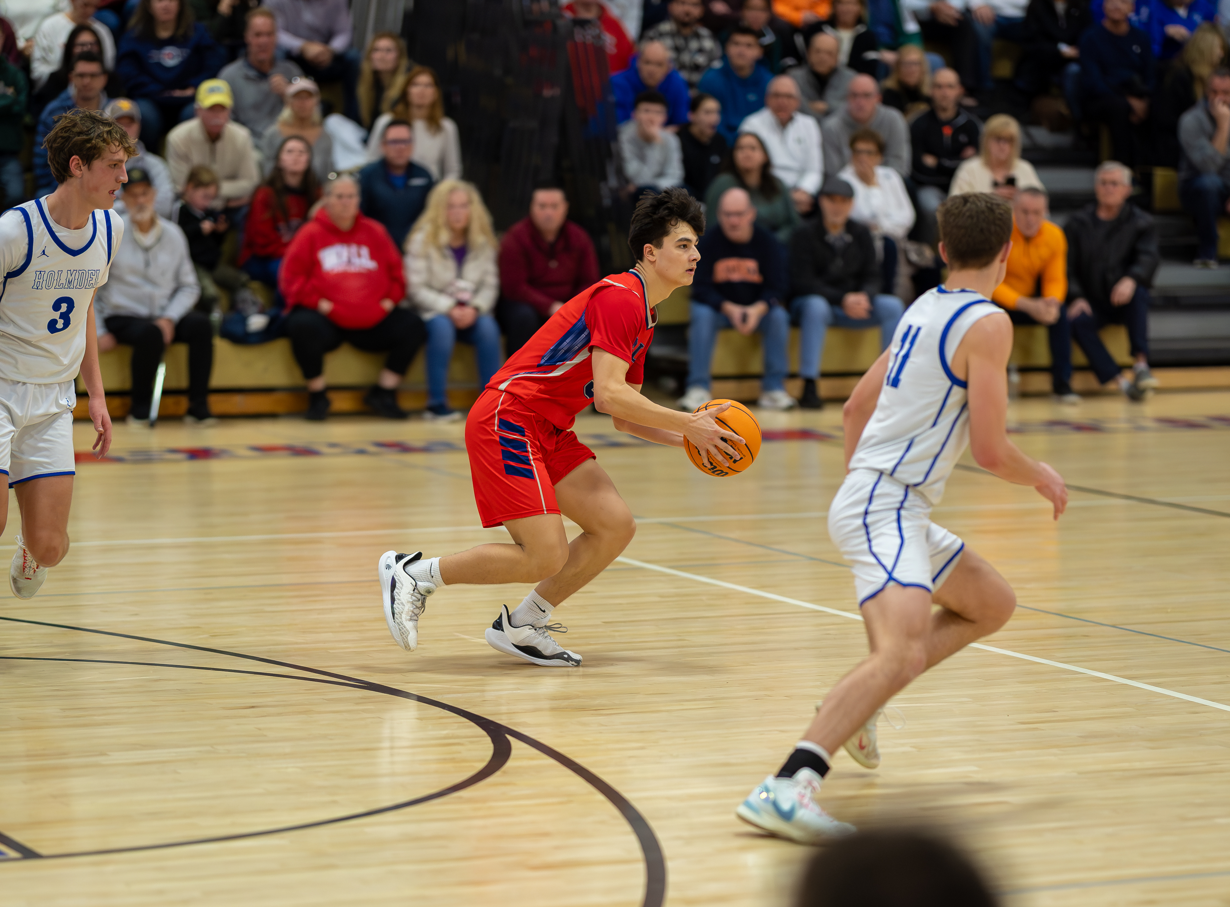 A basketball player in a red jersey dribbles the ball on a wooden court while spectators watch from bleachers in the background.