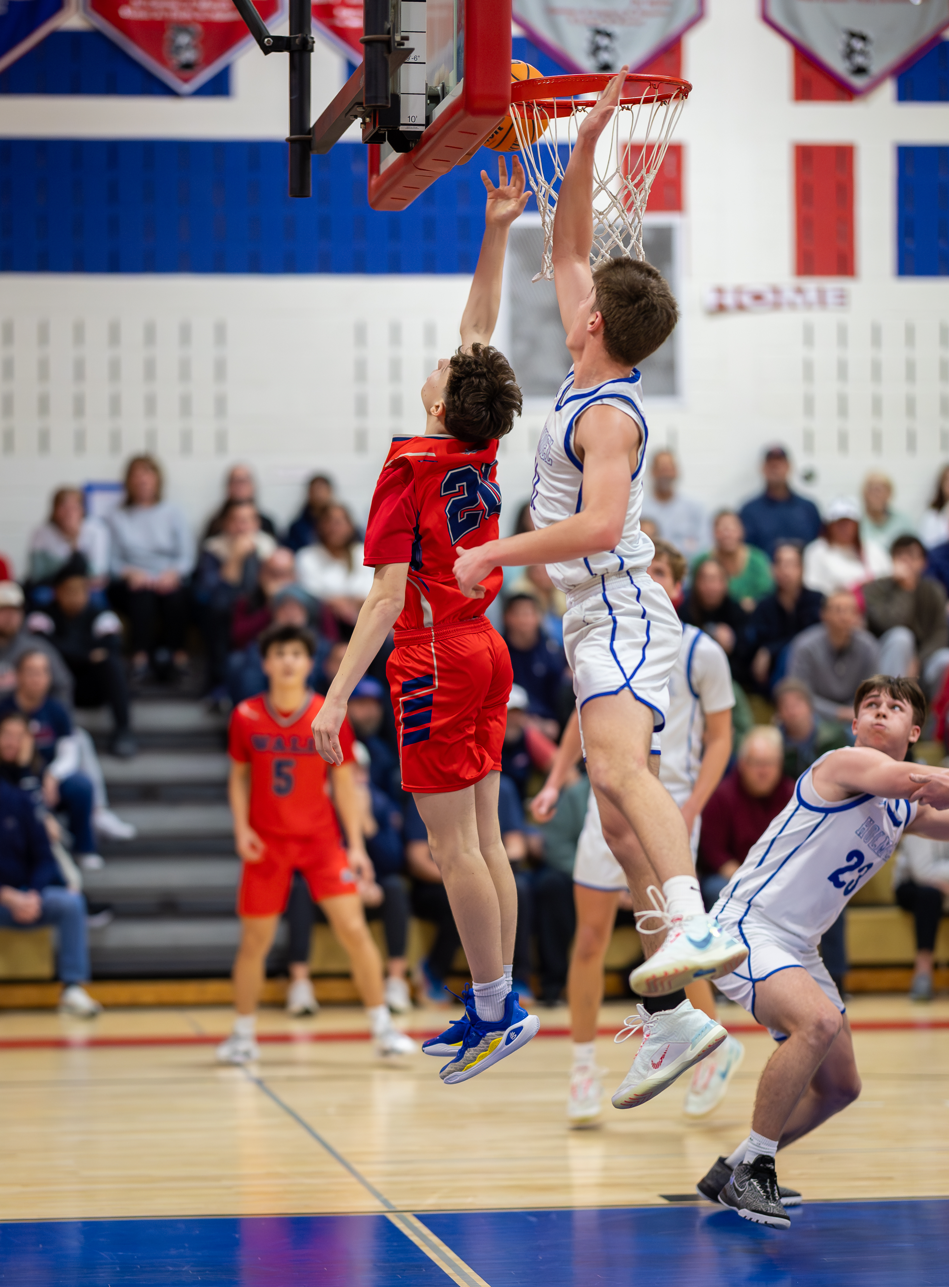 Two basketball players in red and white uniforms jump for a shot at the hoop in the foreground, while spectators fill the bleachers in the background.