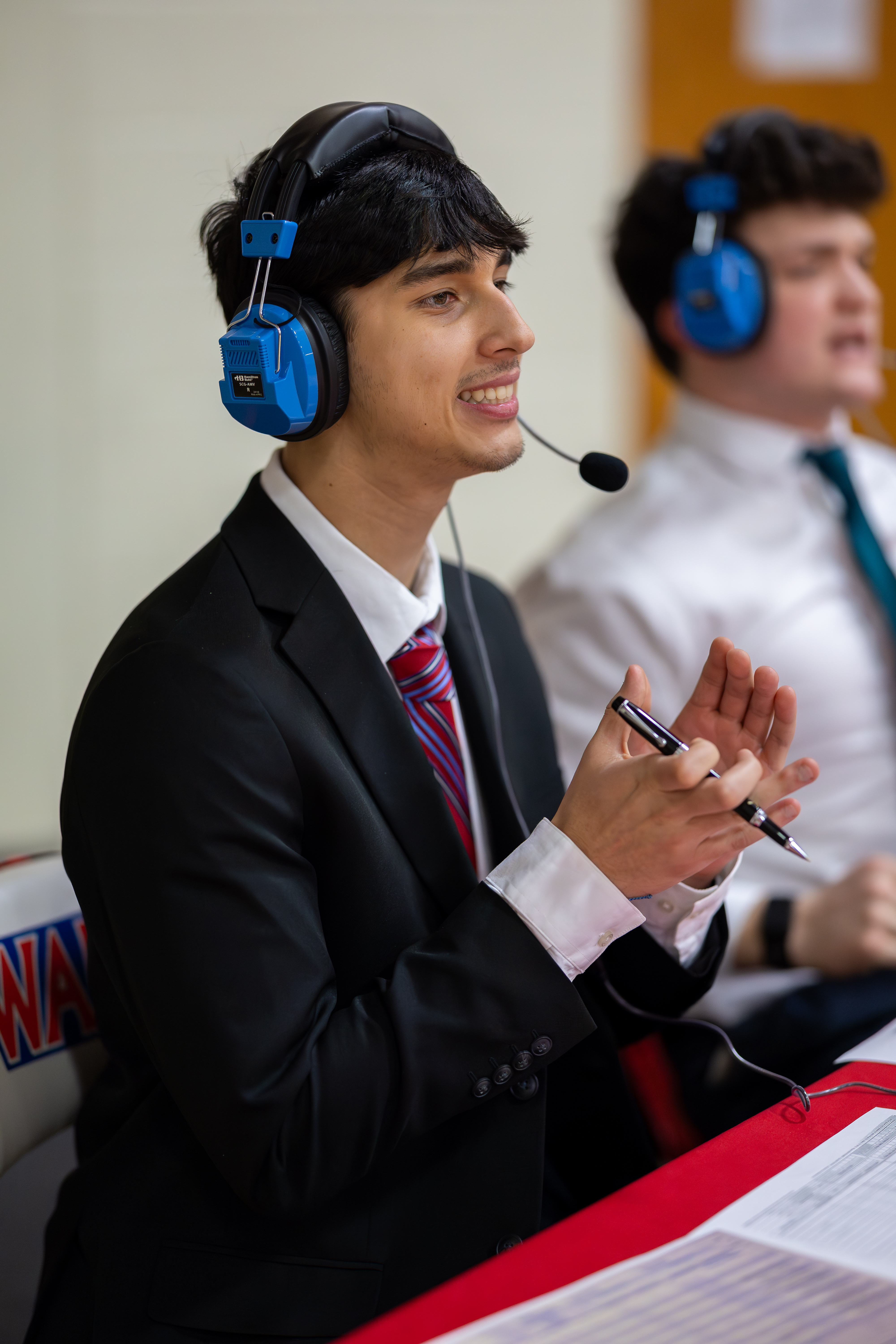 A young man in a suit and headphones speaks into a microphone in the foreground, while another person in similar attire is visible in the blurred background.