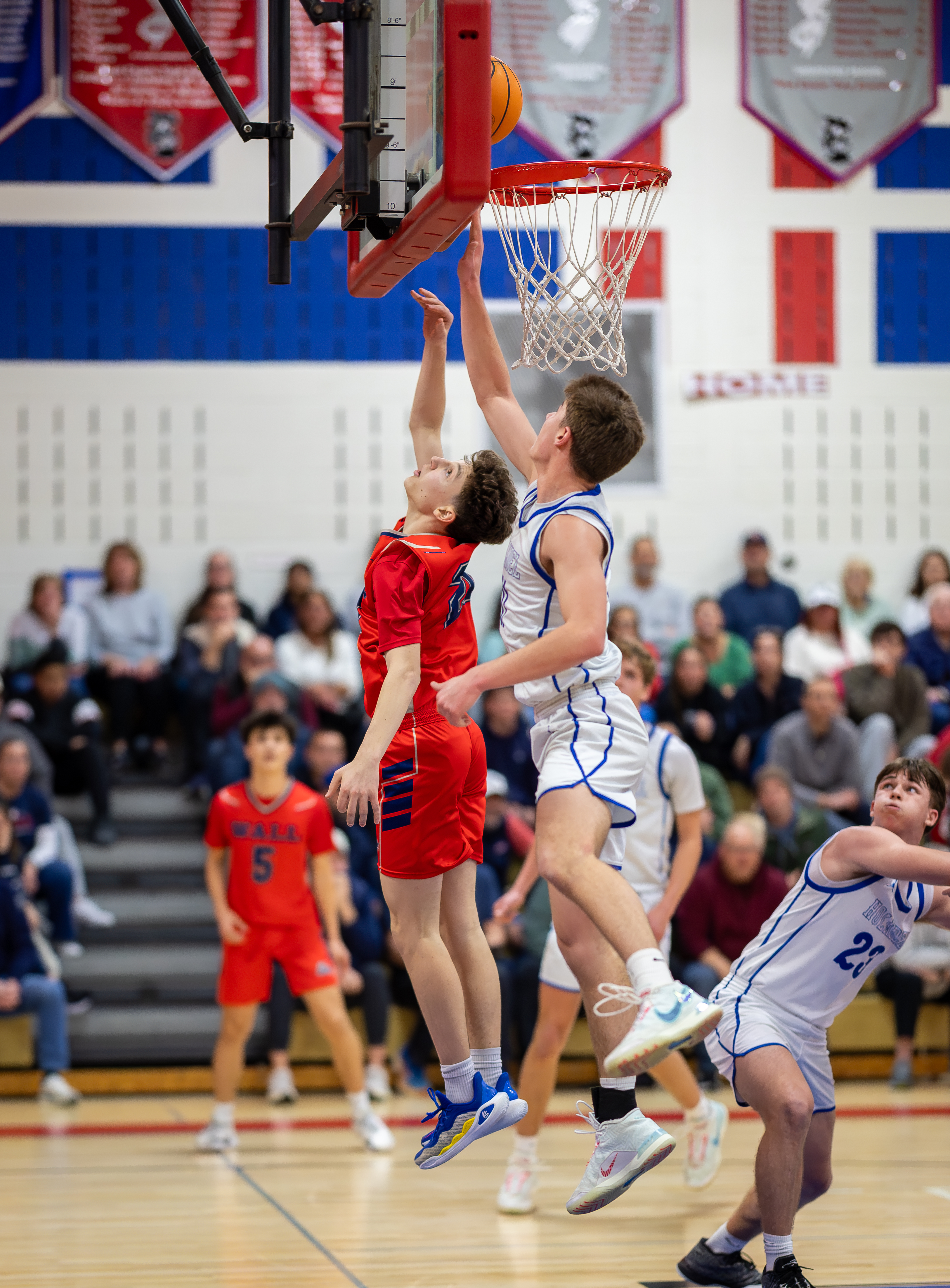 Two basketball players in red and white uniforms jump to score a basket in the foreground, while spectators fill the bleachers in the background.