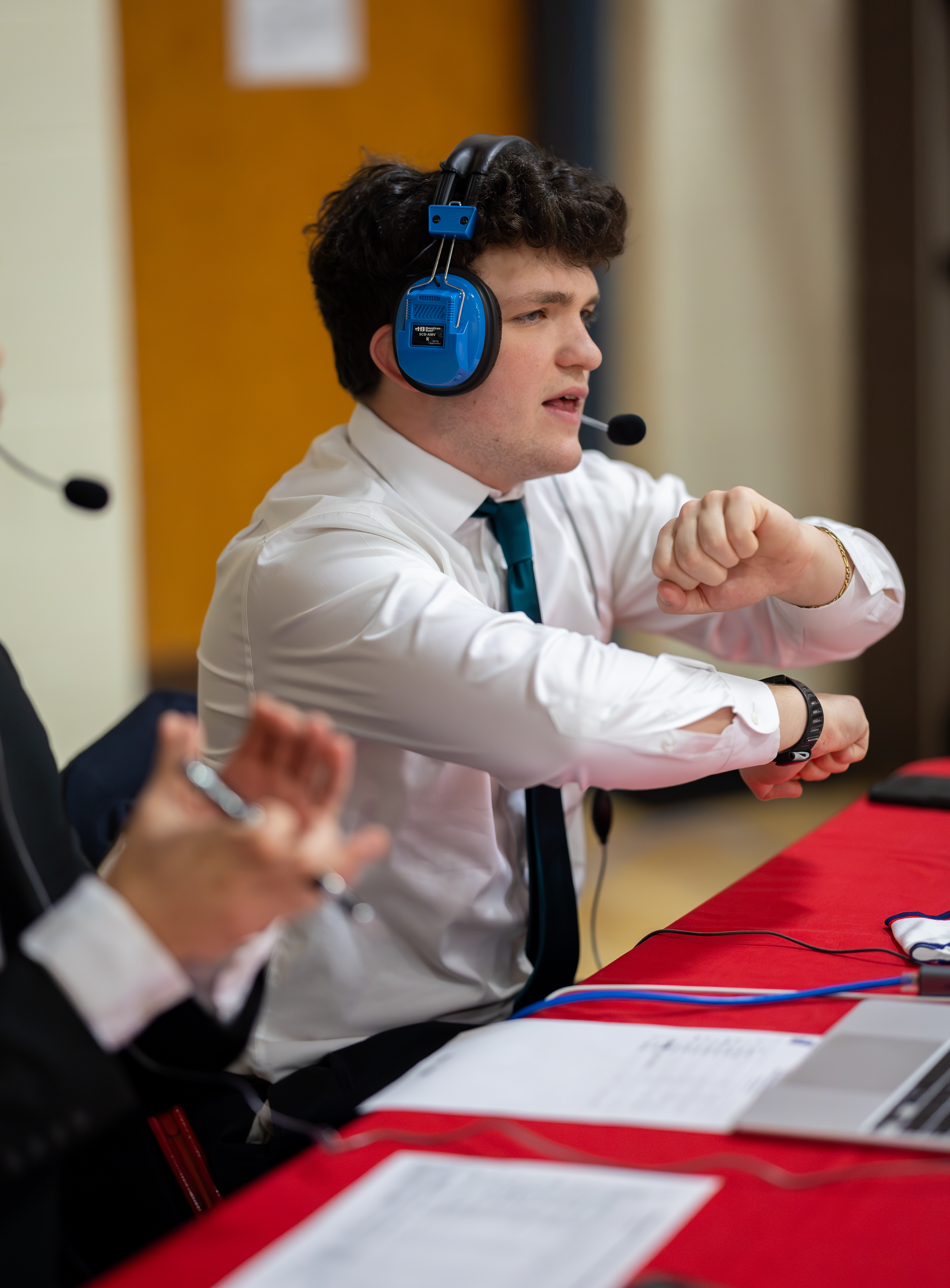 A young man in a white shirt and tie, wearing headphones, gestures with his hand while seated at a red table with a laptop and papers, with a blurred background of a gymnasium.