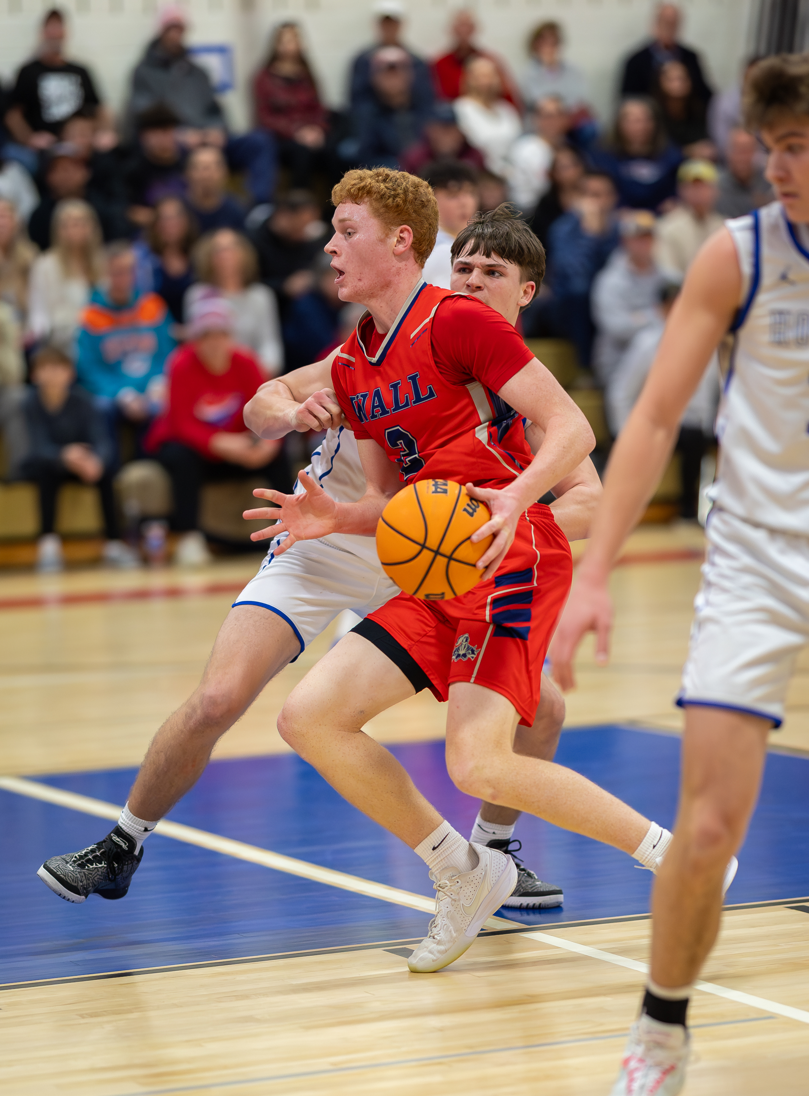 A young basketball player in a red jersey dribbles the ball while being defended by an opponent, with a blurred crowd watching from the background.