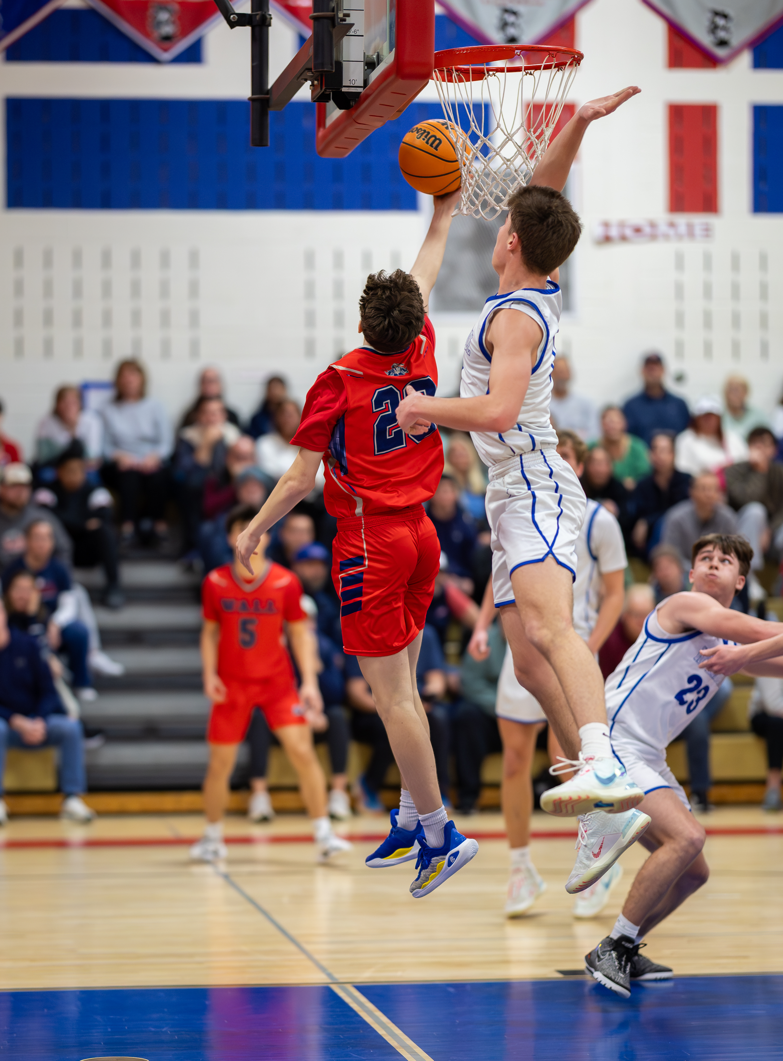 Two basketball players in red and white uniforms leap for a rebound in the foreground, while spectators fill the bleachers in the background.