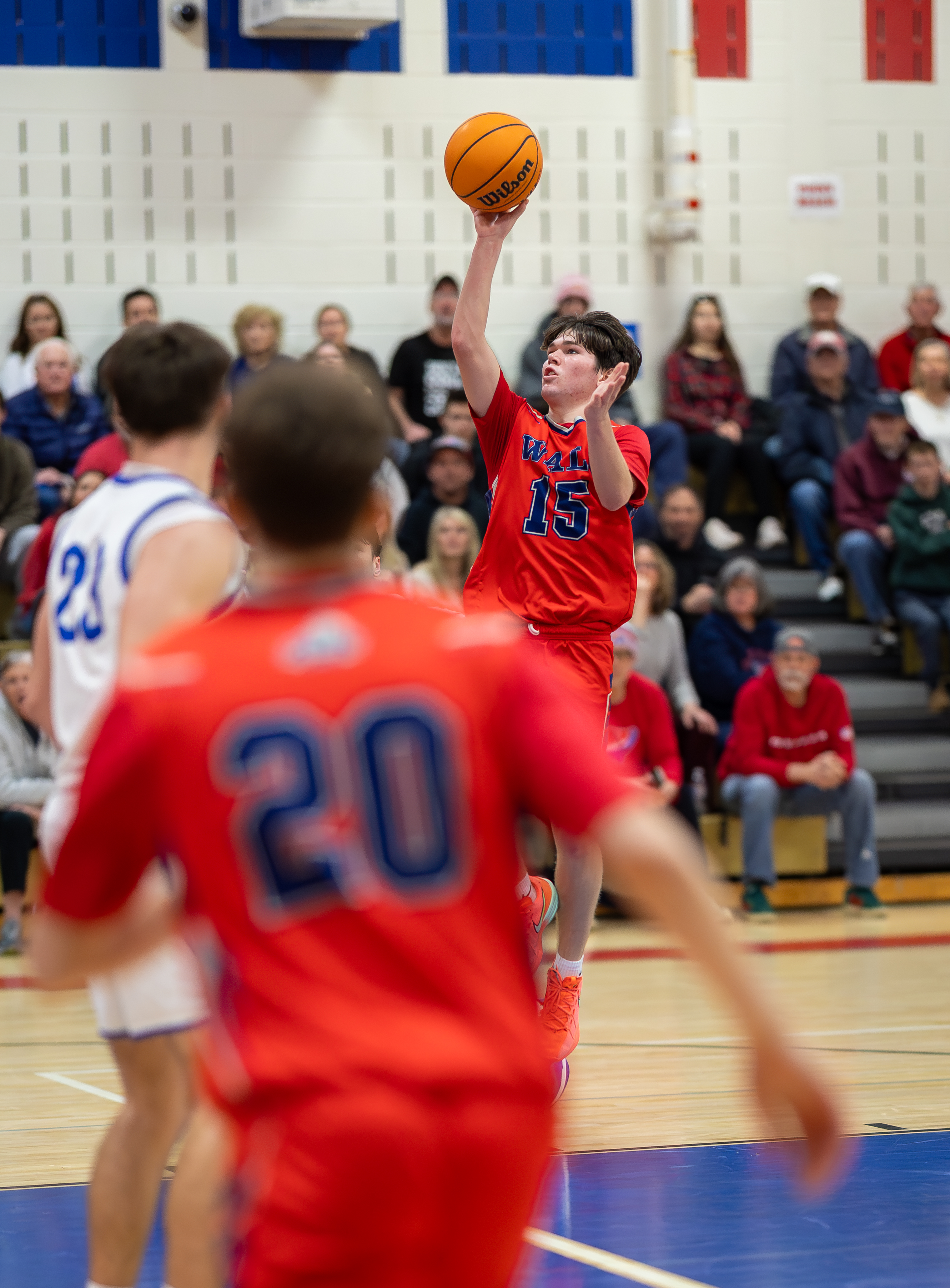 A basketball player in a red jersey with the number 15 on it shoots a basketball, with blurred players in red and blue jerseys in the foreground and spectators in the background.