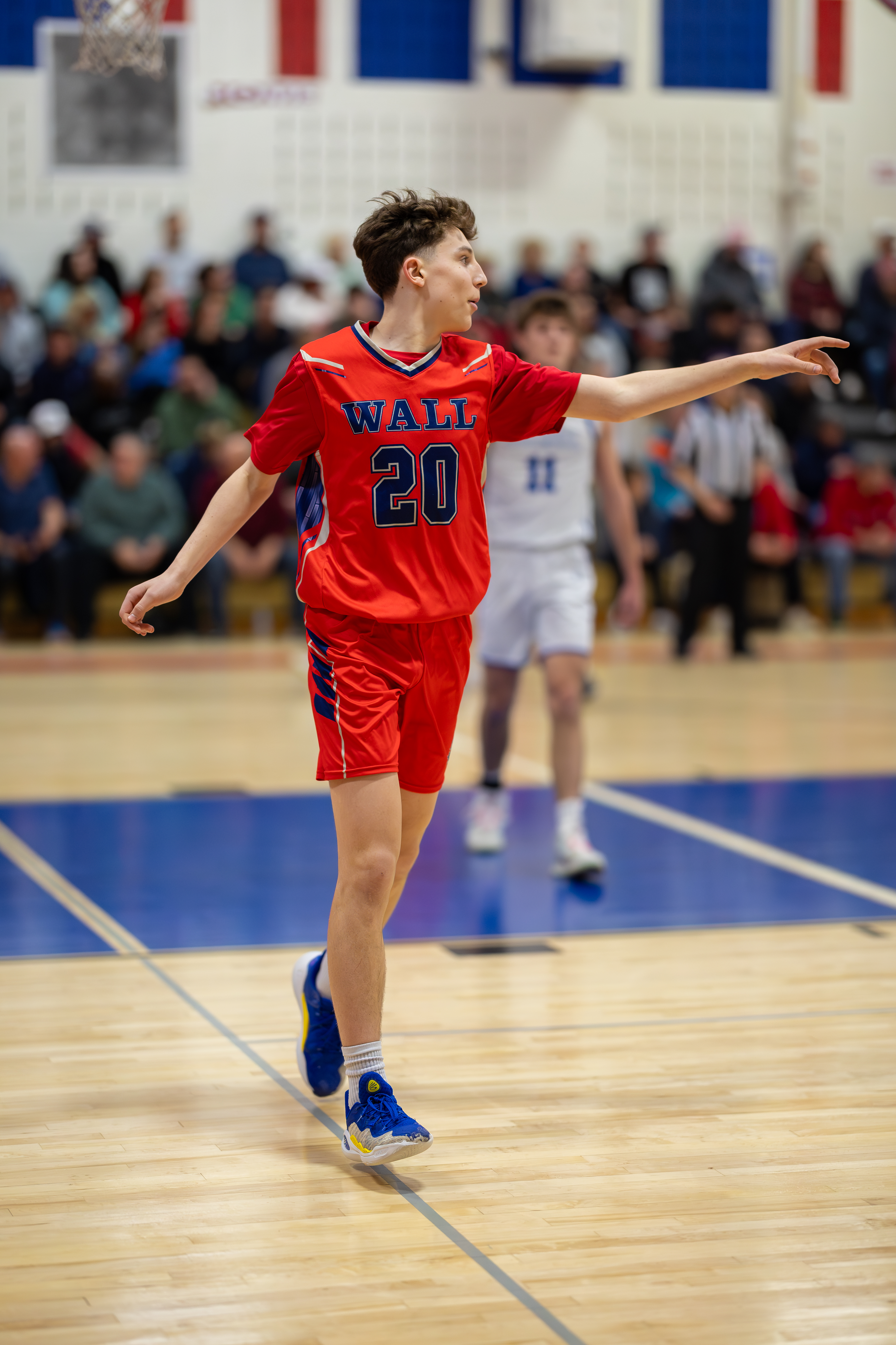A young basketball player in a red uniform with the number 20 on it stands on a blue court, with spectators in the background.