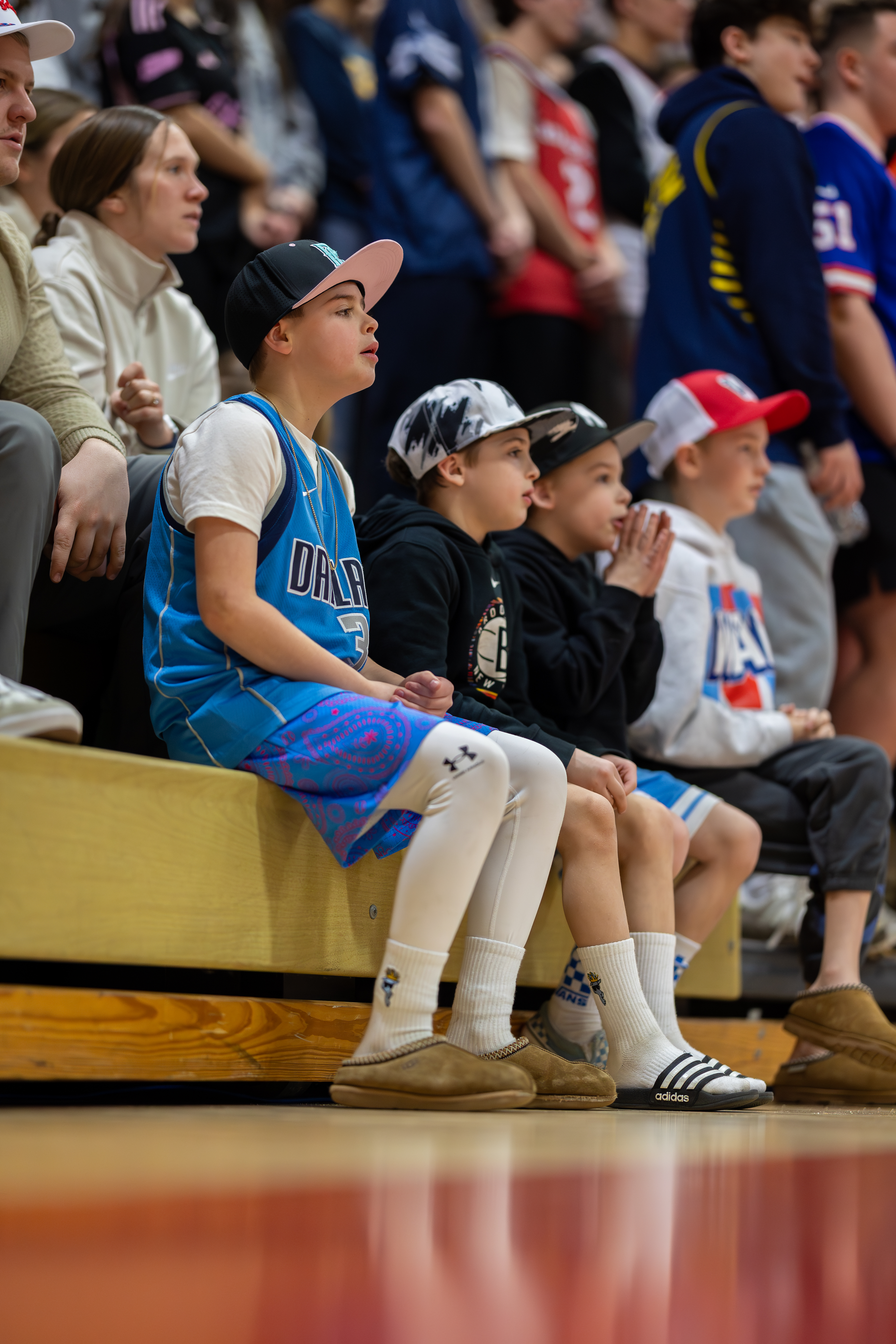 Young boys in casual attire sit in the foreground watching a sporting event, while a blurred crowd of spectators fills the background.