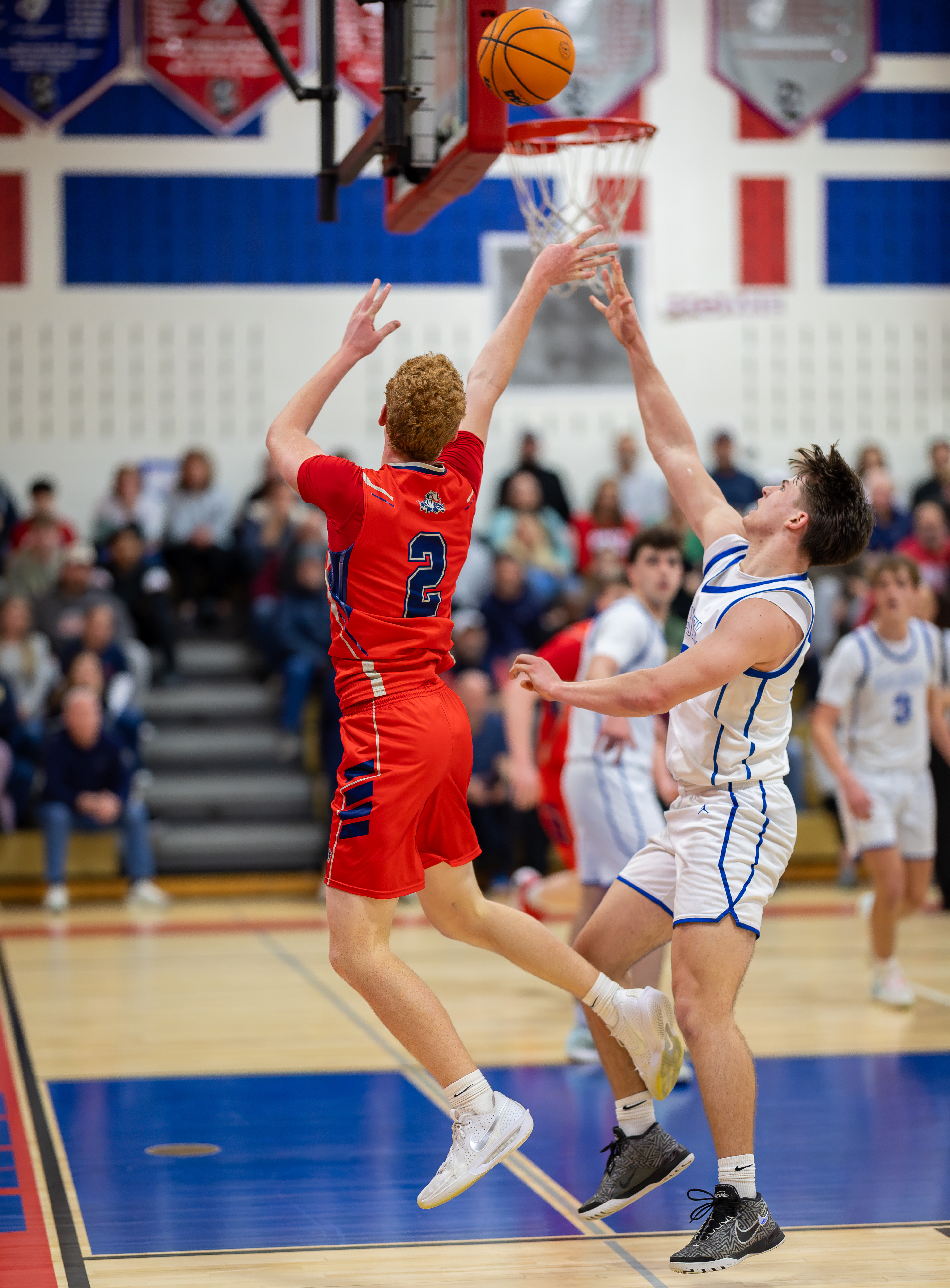 Two basketball players in red and white uniforms leap for a shot in the foreground, with a blurred crowd watching from the bleachers in the background.