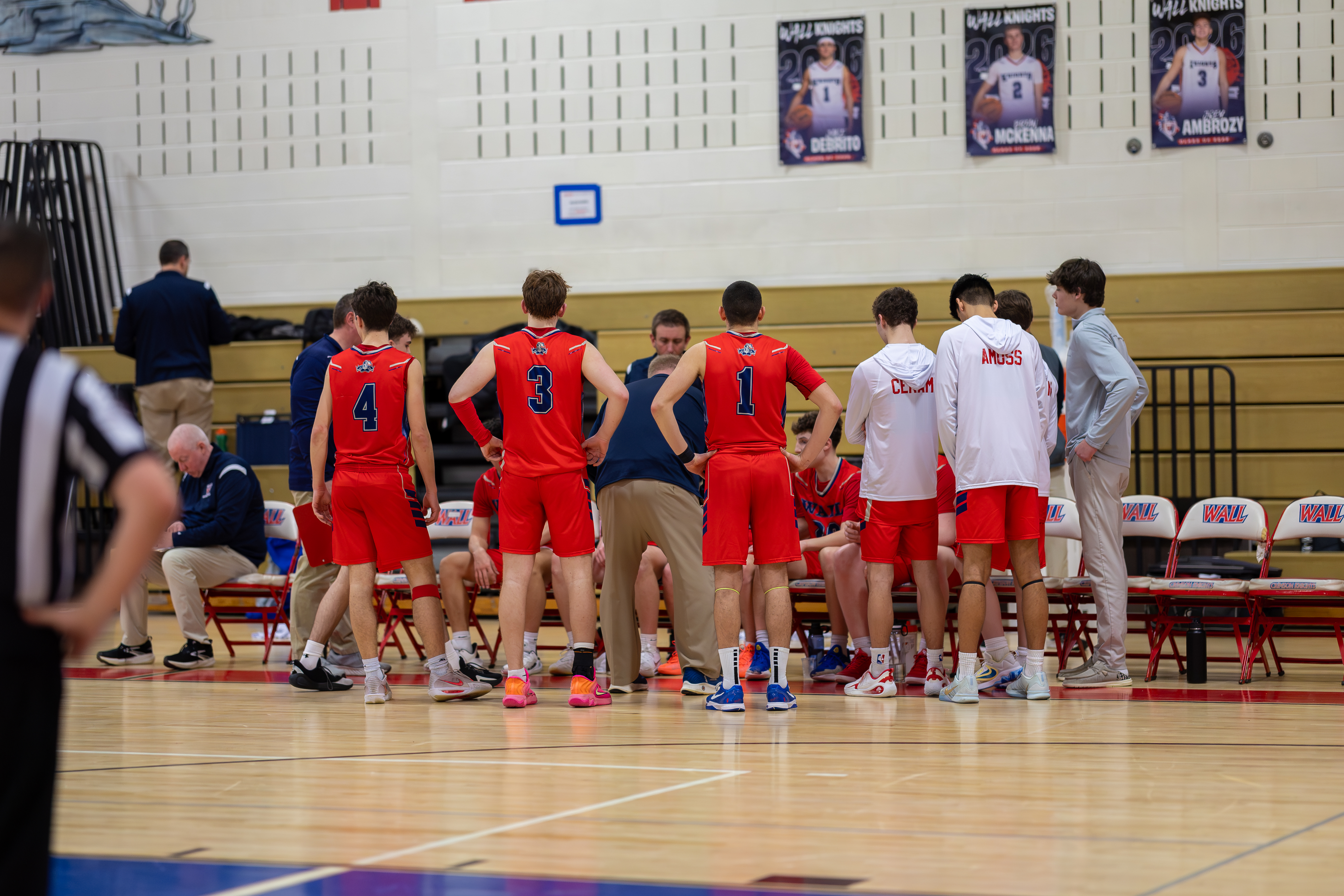 A basketball team in red uniforms huddles around their coach on the court, with posters of players on the wall in the background.